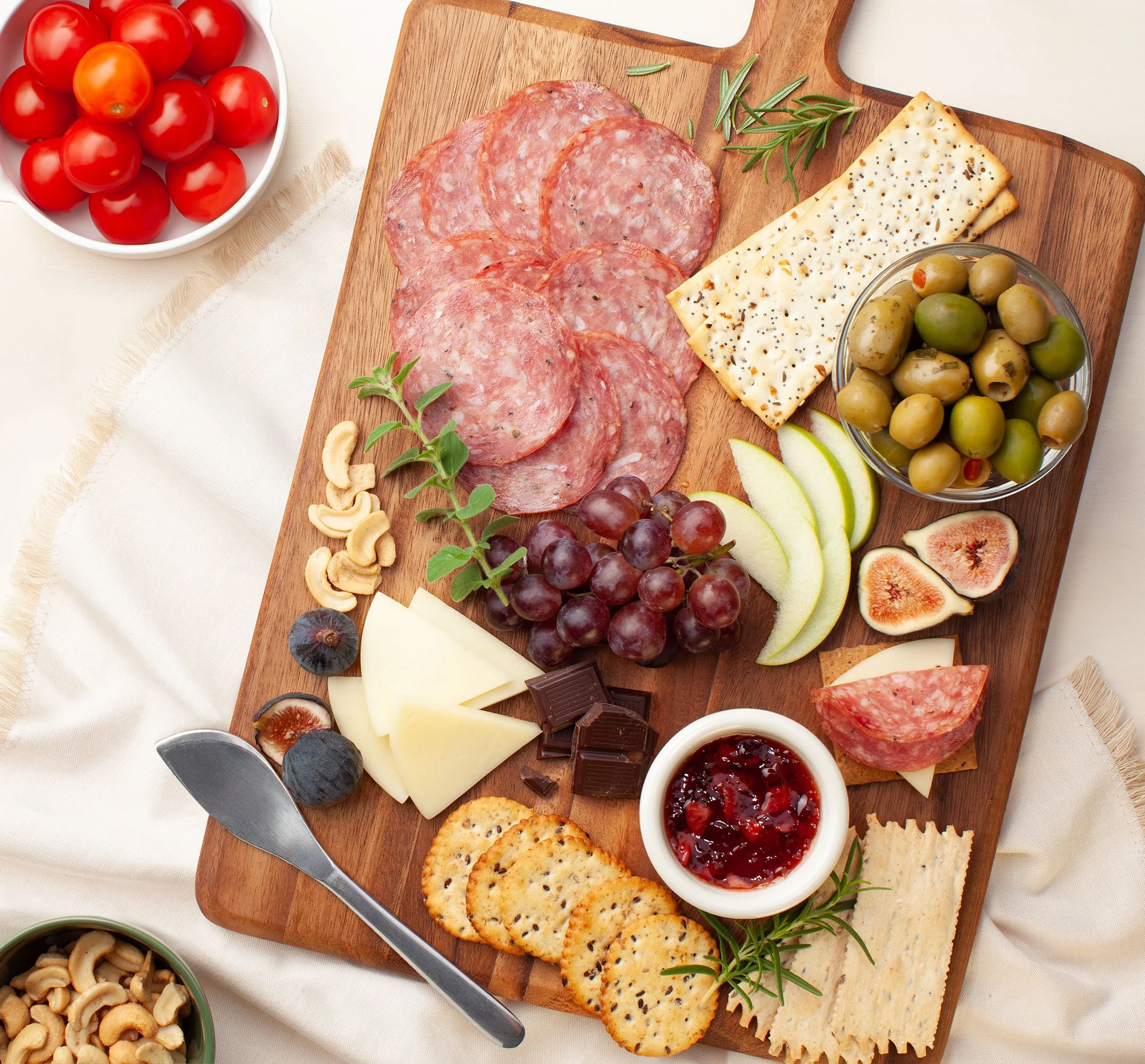 Charcuterie board with salami, cheese, crackers, olives, grapes, figs, pears, tomatoes, nuts, rosemary, and fruit preserves