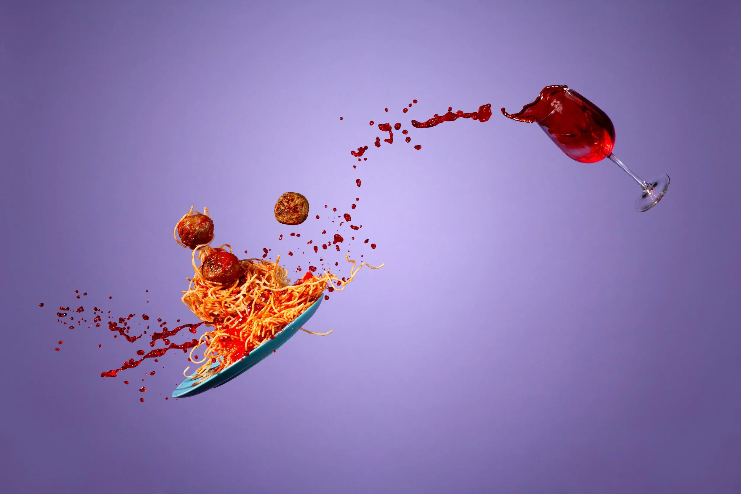 Spaghetti and meatballs splashing from a blue plate against a tipped glass of red wine on a purple background