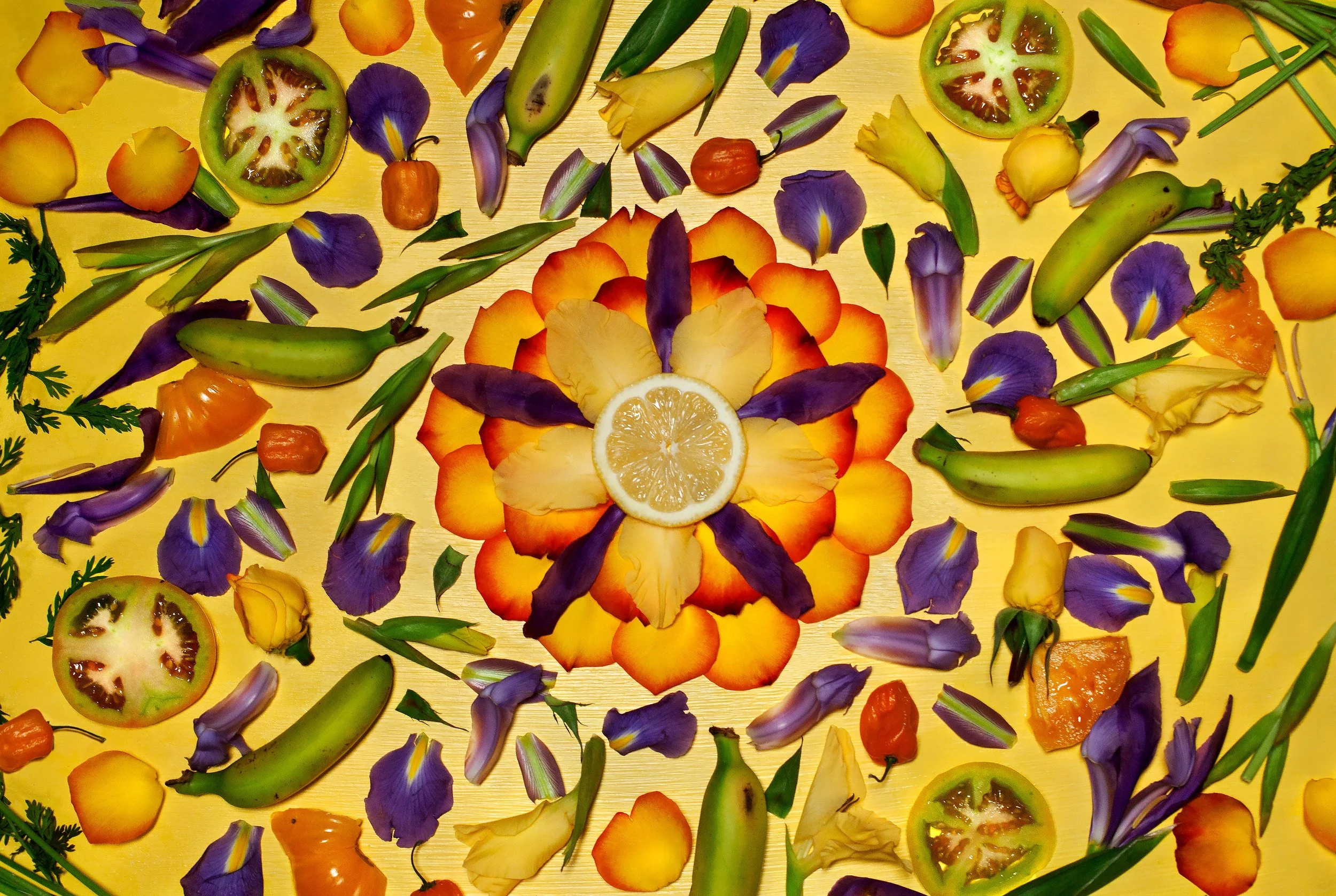 Overhead design of colorful edible flowers, green tomatoes, peppers, bananas, herbs, and a lemon slice in a kaleidoscope arrangement on a yellow background