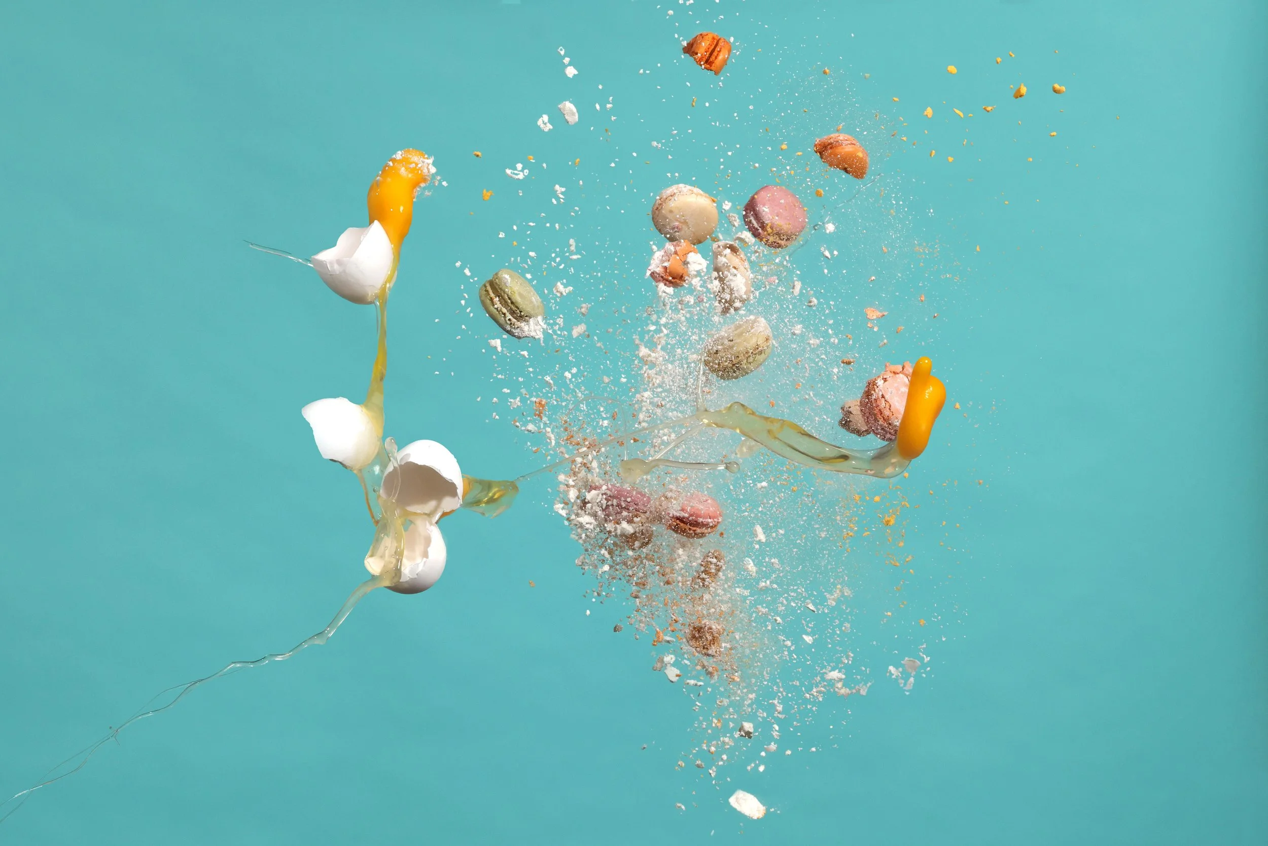 Colorful macarons, eggs, flour, and baking ingredients splashing and colliding against a turquoise background
