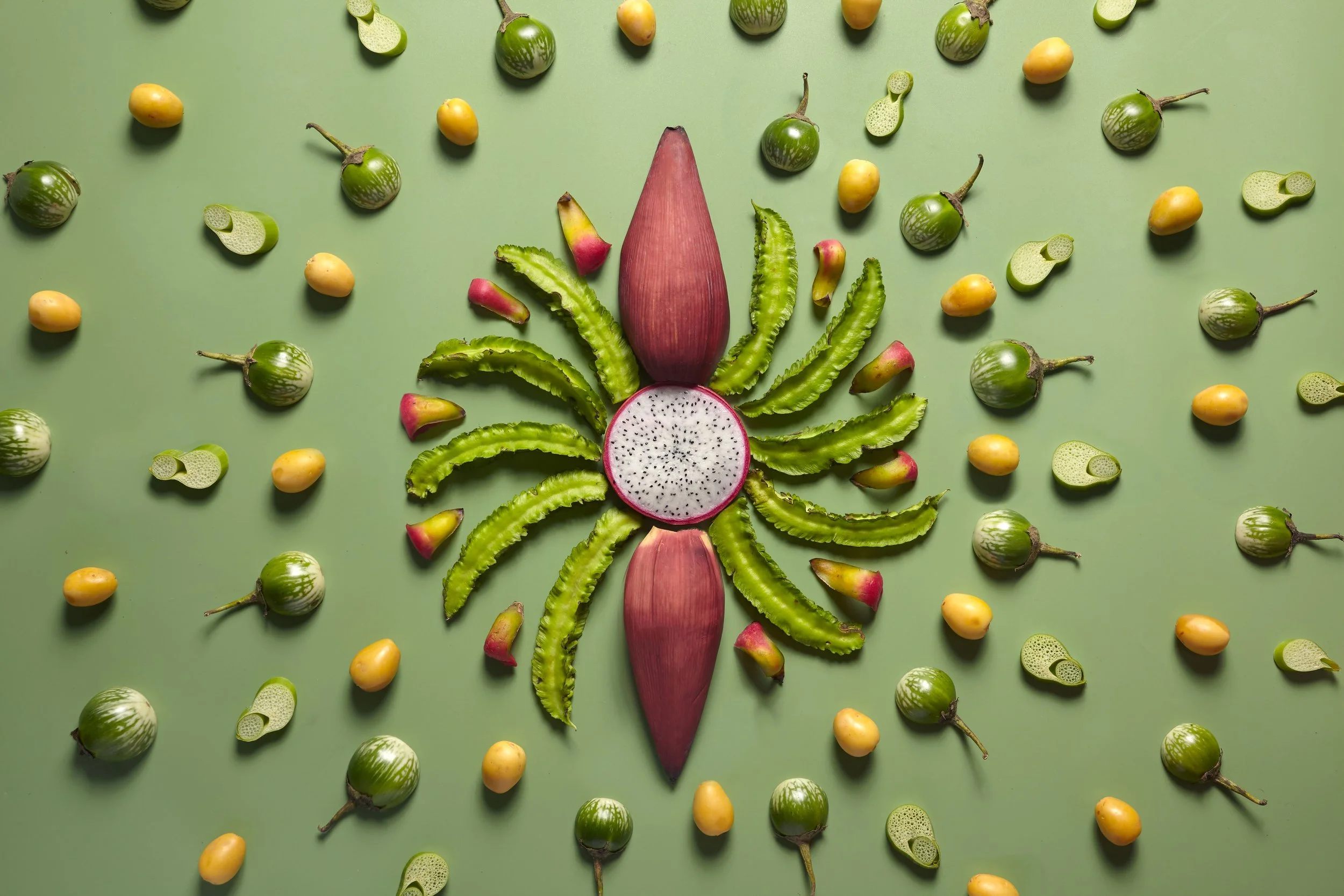 Overhead arrangement of dragon fruit, winged beans, banana blossoms, yellow fruits, and striped green eggplants on a green background