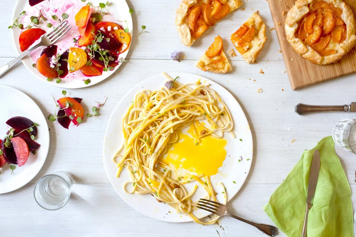 Overhead view of linguine with yellow sauce, beet and citrus salad, and apricot galette on a rustic table