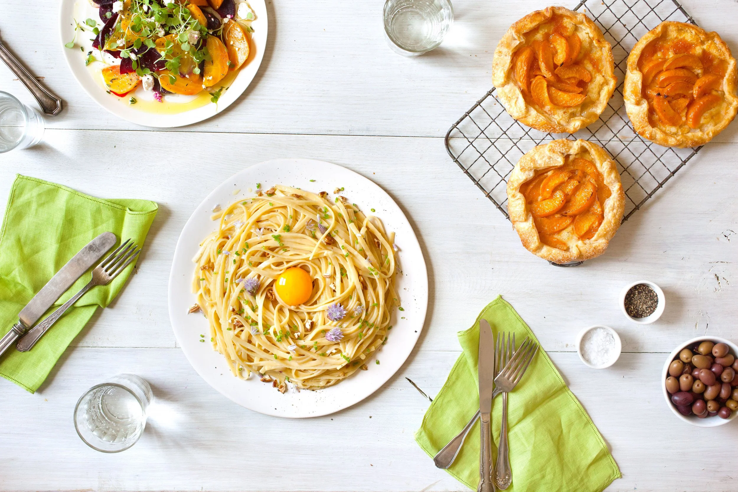 Overhead view of a colorful meal with creamy pasta, roasted beet and carrot salad, apricot galettes, green napkins, olives, and tableware on a white table.