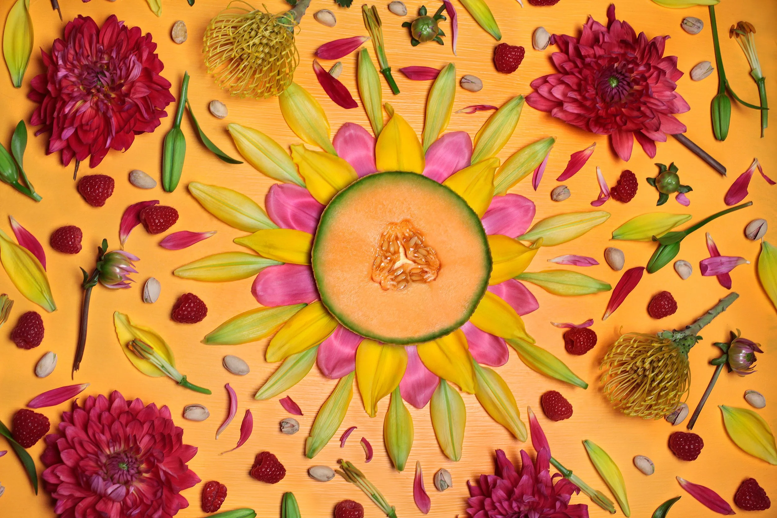 Overhead arrangement of a sliced cantaloupe surrounded by colorful flowers, petals, raspberries, seeds, and plant stems on an orange background
