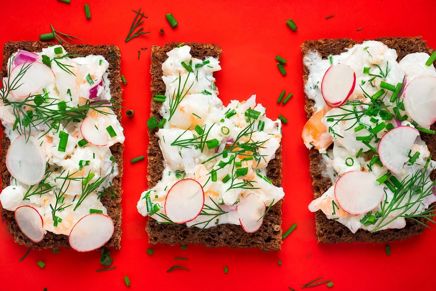 Three open-faced rye sandwiches topped with creamy salad, smoked salmon, radishes, dill, and chives on a red background