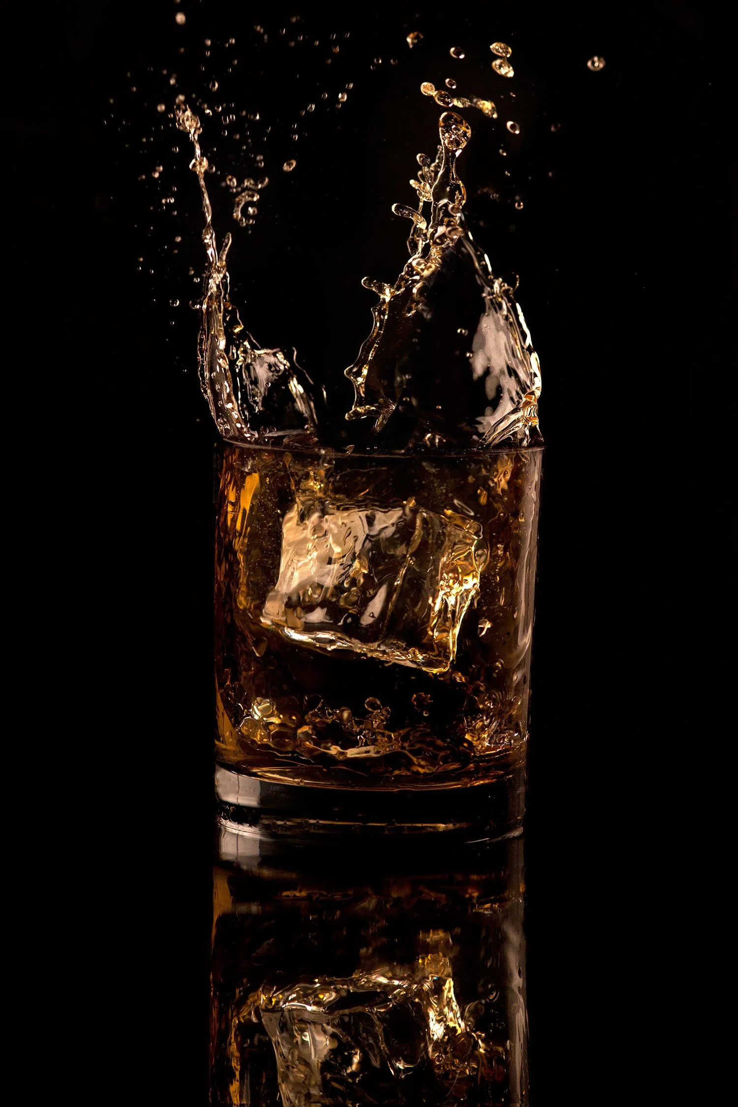 Close-up of amber whiskey splashing in a rocks glass against a black background and reflective black tabletop surface