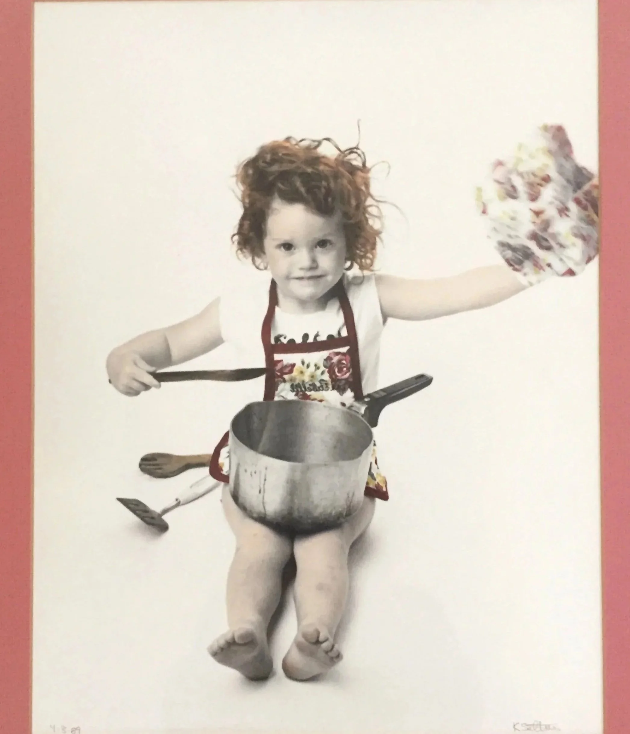 Child in a floral apron holding a wooden spoon beside a large cooking pot and utensils.