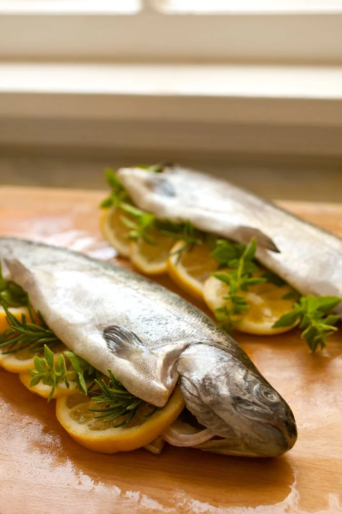 Two whole silver fish garnished with lemon slices and fresh herbs on a wooden cutting board