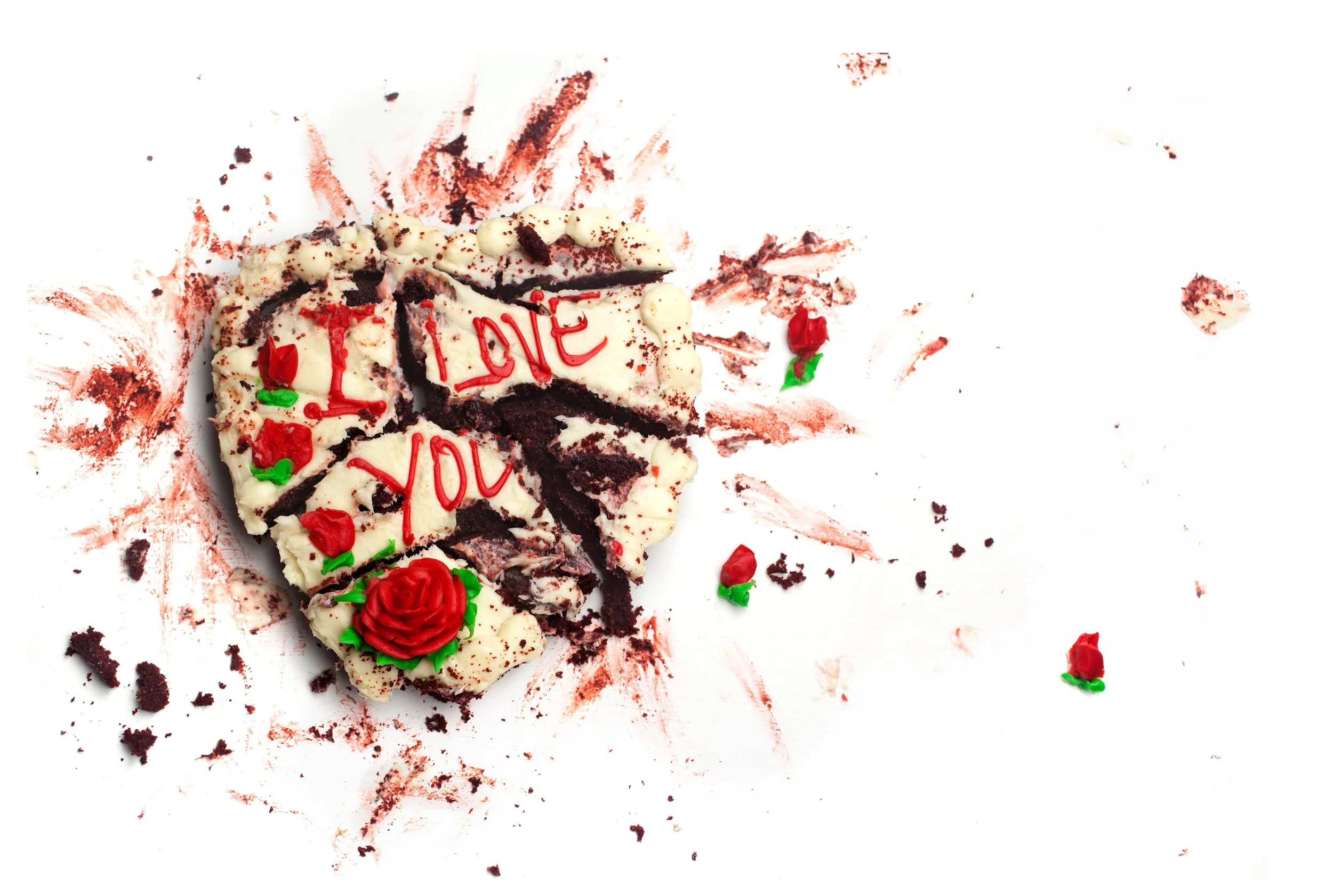 Messy partially cut up chocolate cake that says I Love You, with white frosting, red icing, and decorative roses on a white surface