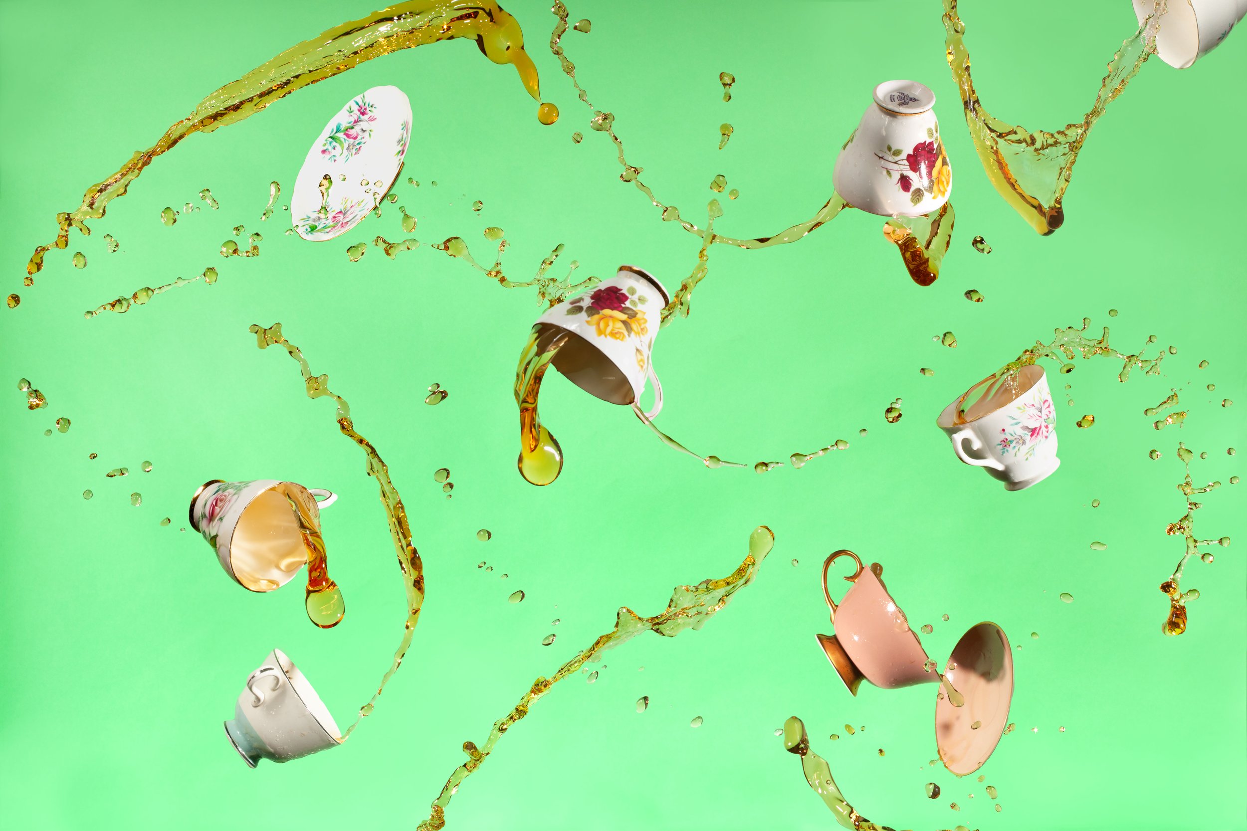 Floral porcelain teacups and saucers splashing through amber tea against a bright green background