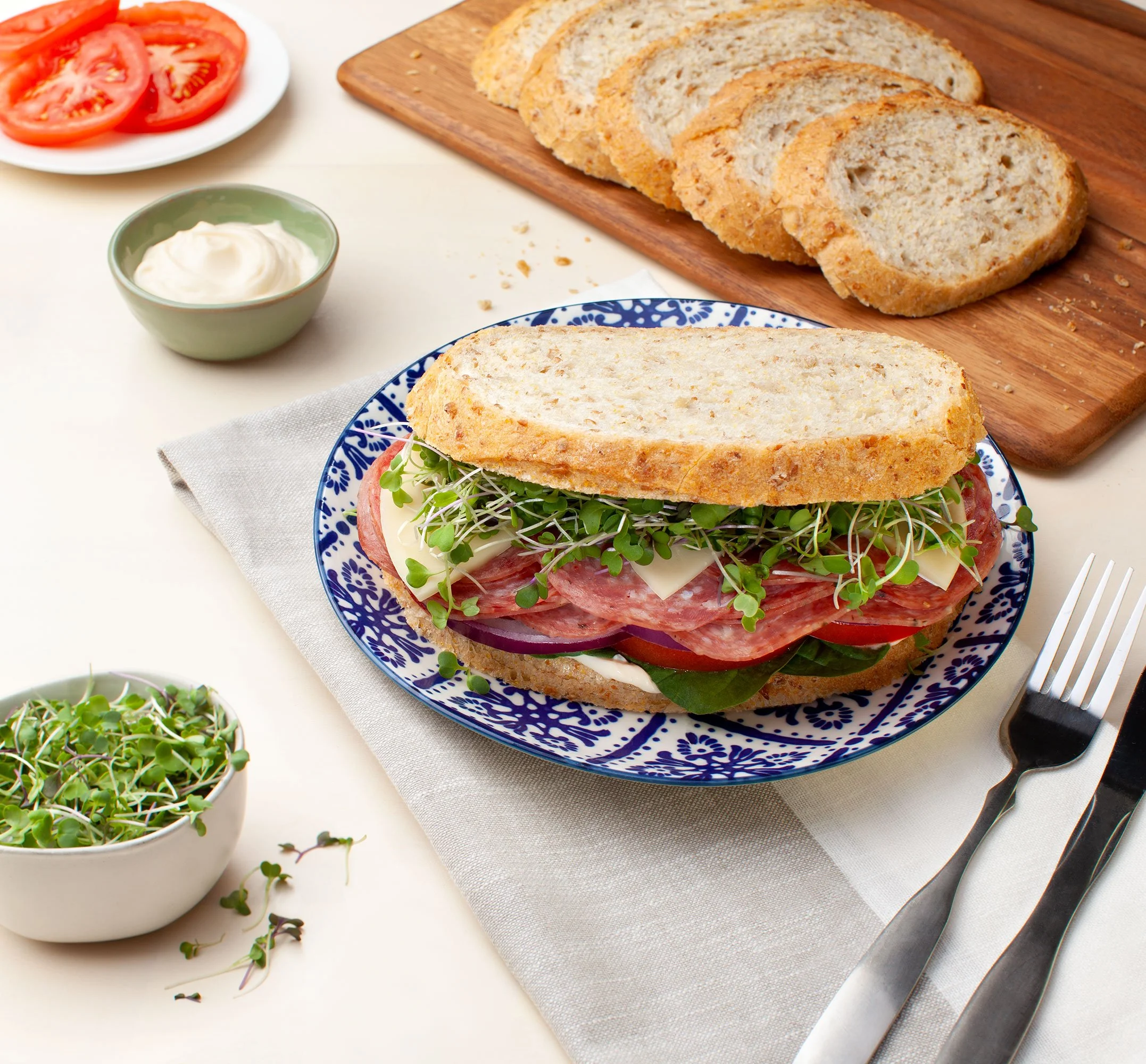 Gourmet sandwich with salami, cheese, tomato, spinach, and microgreens on multigrain bread, served with sliced tomatoes and mayonnaise