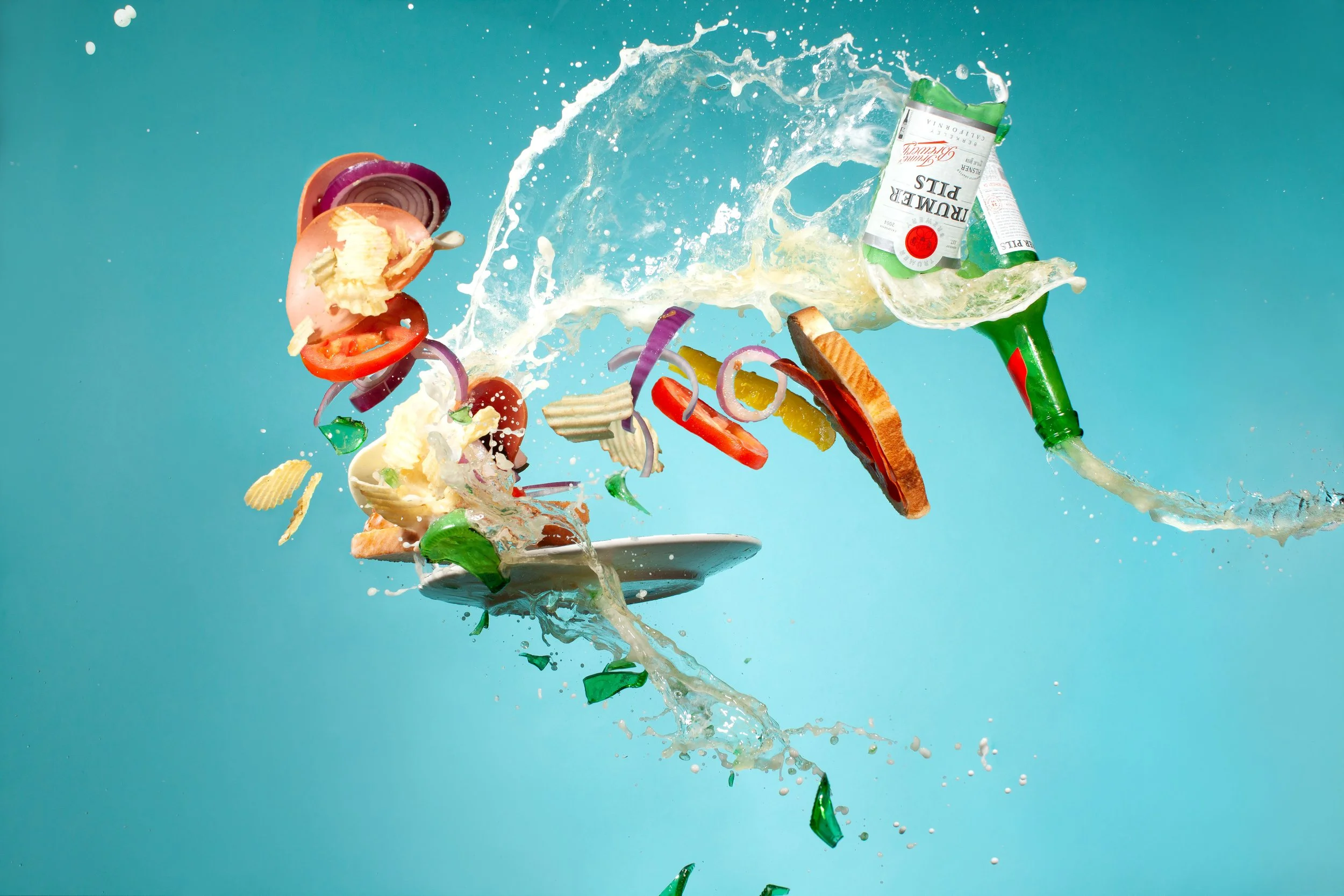 Bottle of beer breaking against a sandwich plate mid air. Sandwich ingredients (pickels, tomato, red onion, meat) and potato chips are flying through the giant splash of liquid beer, all against a turquoise background