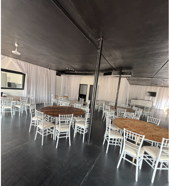 Event space with round wooden tables and white chairs, black ceiling, white curtains, and large windows. essential or deluxe package uncovered tables