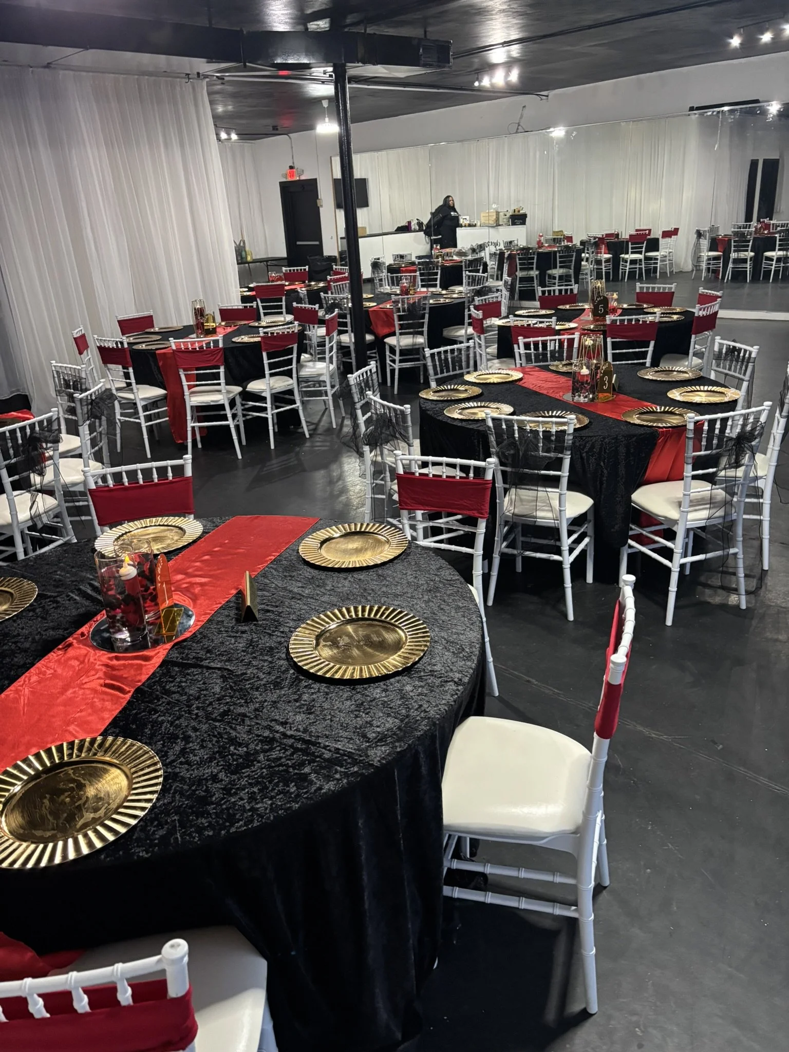 Elite Banquets event space decorated for a celebration with round tables covered in black tablecloths, red table runners, gold paper plates, and black and red chairs with some black bows. fully dressed celebration
