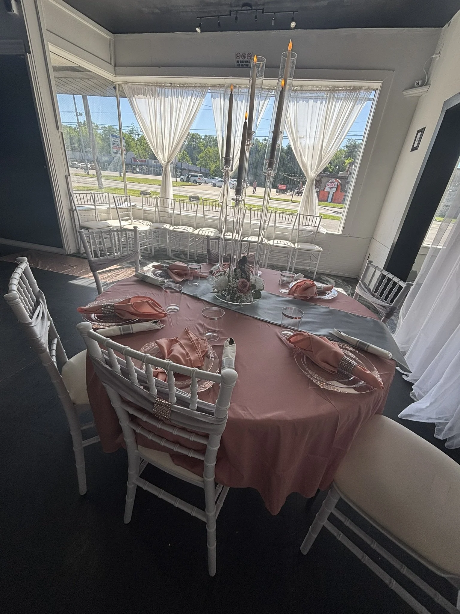 Elegant dining setup with a round table covered with a pink tablecloth, white chairs arranged around it, pink napkins and glasses set on the table, and a floral centerpiece featuring roses and candles. Large window with white curtains lets in natural light, showing a street scene outside.