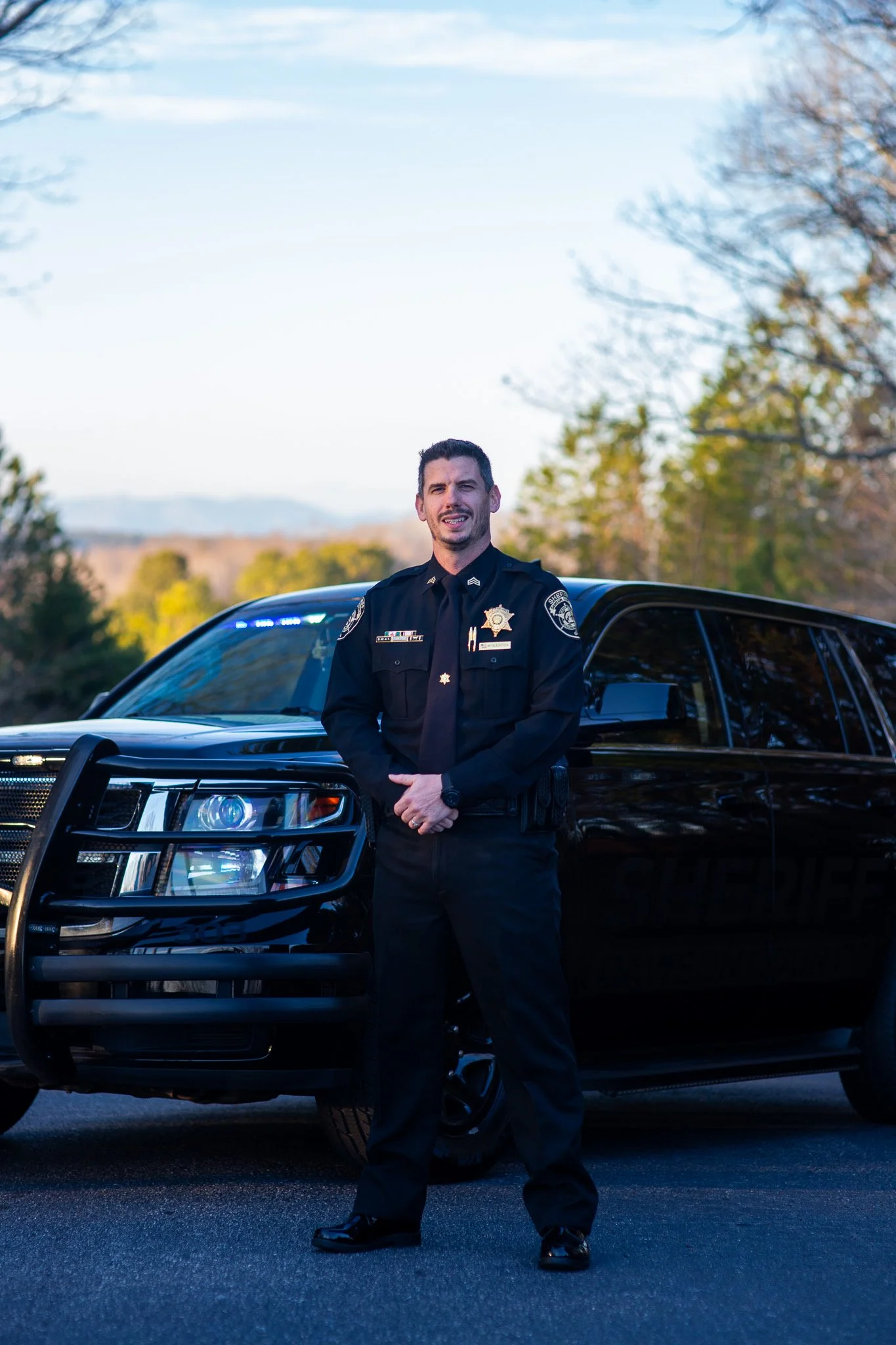 A police officer standing in front of a black police vehicle outdoors, with trees and a clear sky in the background.
