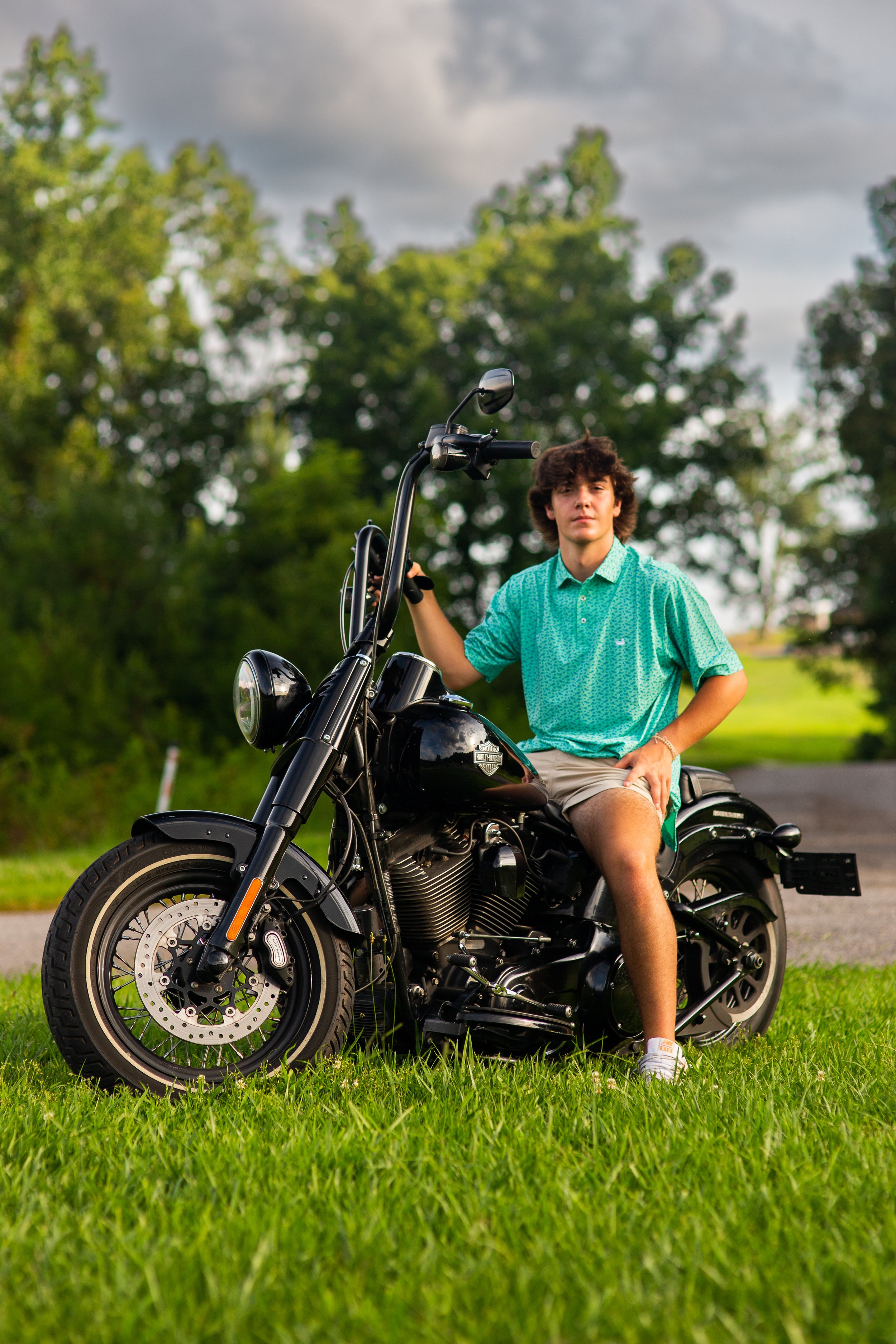 A young man sitting on a black motorcycle in a grassy park area with trees and cloudy sky in the background.