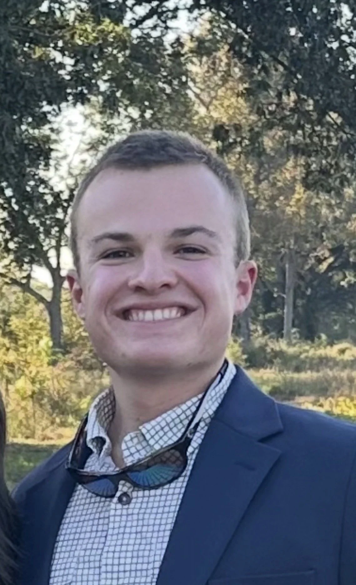 A young man smiling outdoors in a suit jacket and checkered shirt, with trees and sunlight in the background.