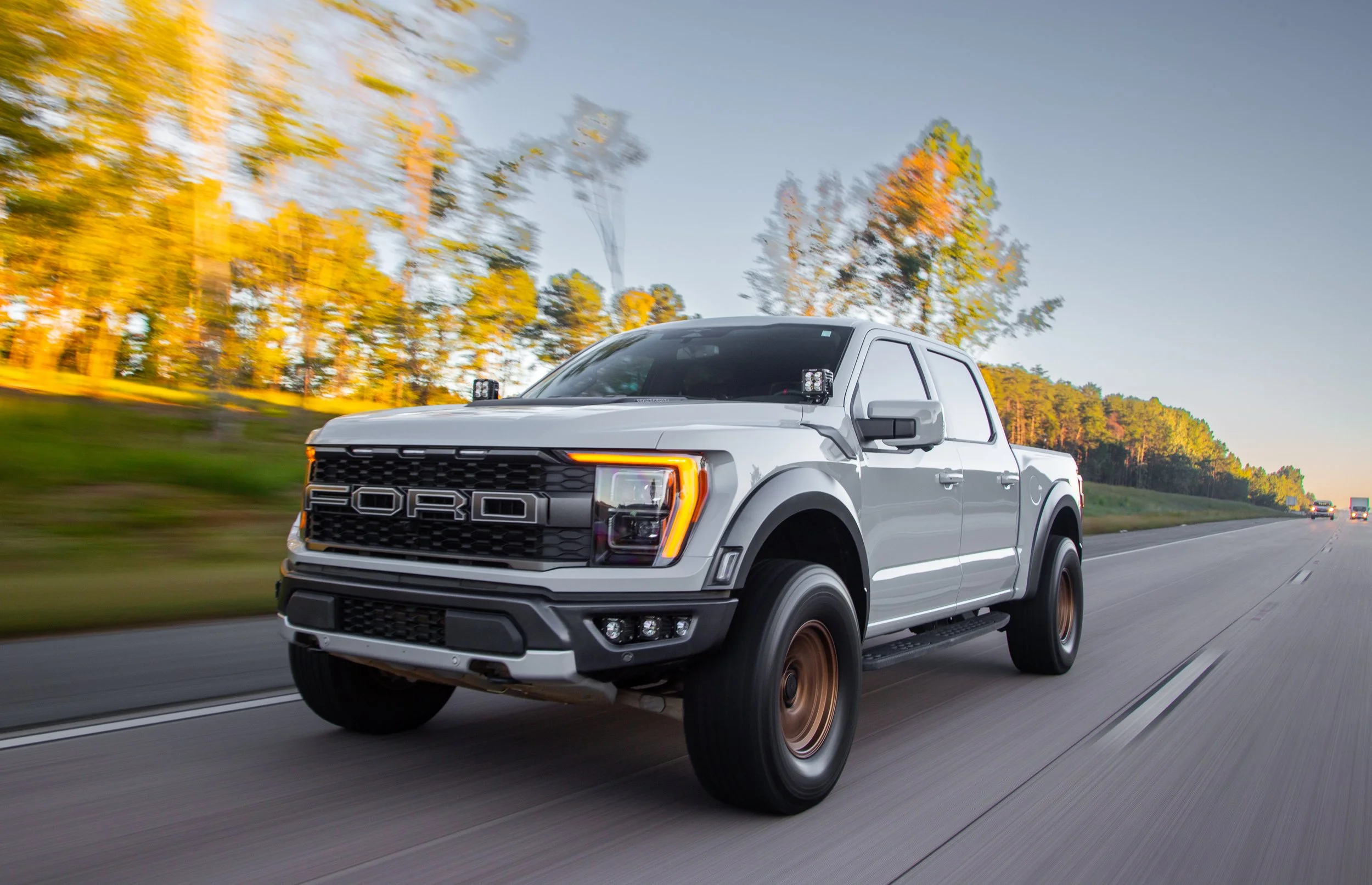A white Ford pickup truck driving on a highway with blurred trees and a blue sky in the background.
