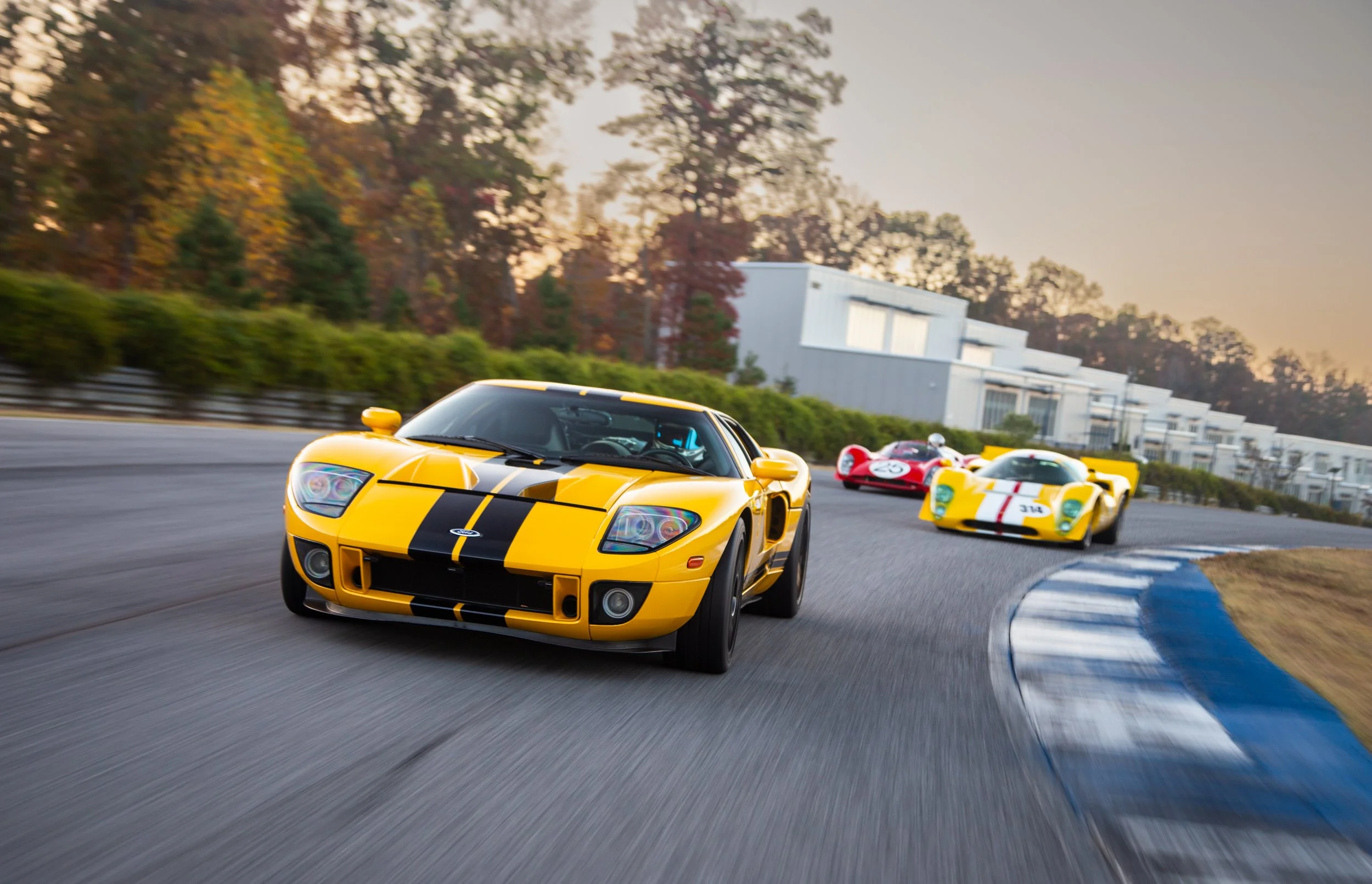 Three race cars on a track during a race, with a yellow car leading and a red car and another yellow car following, on a winding track with trees and buildings in the background at sunset.