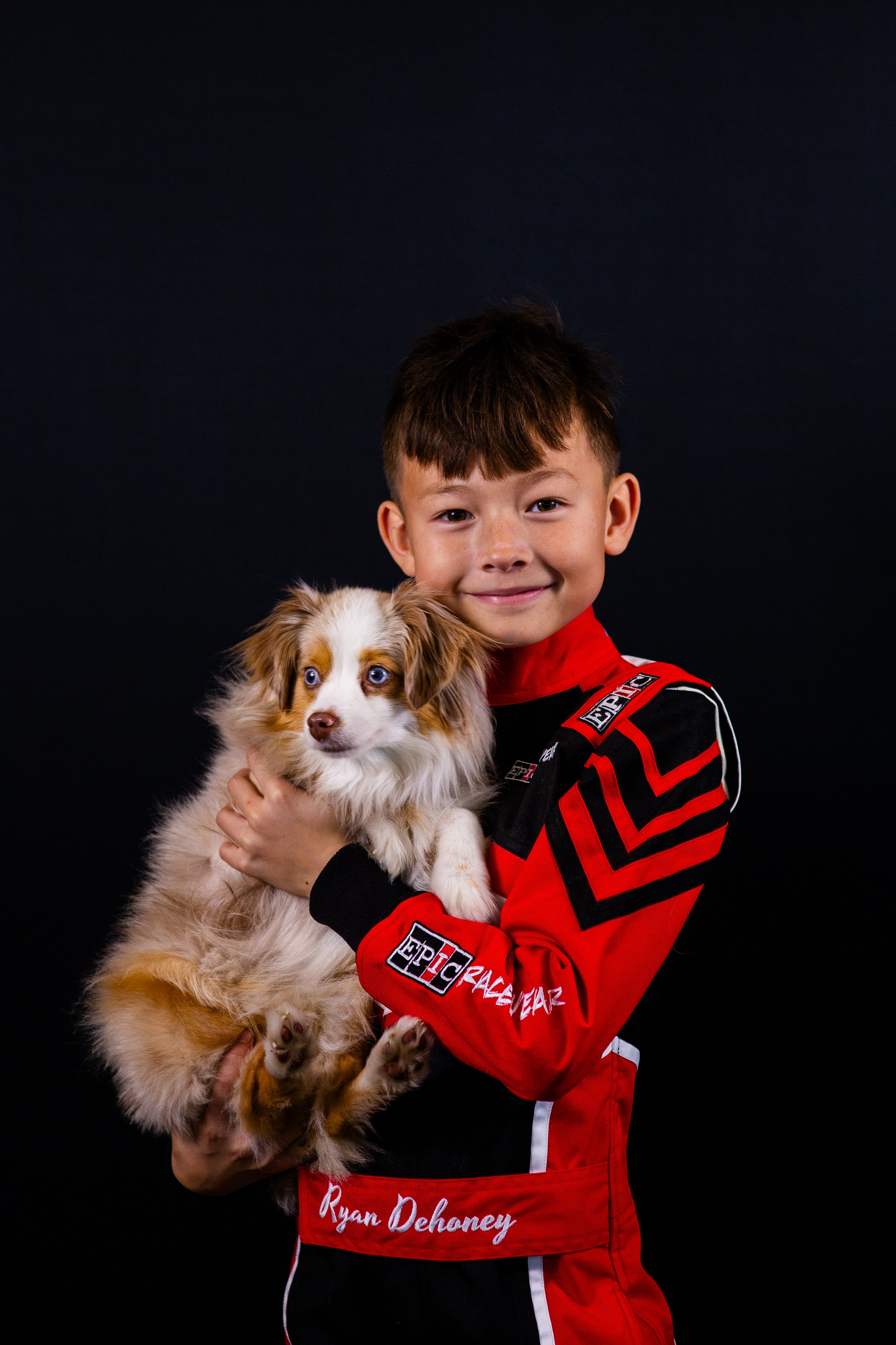 A young boy in a red racing suit holding a fluffy Australian Shepherd puppy with blue eyes against a black background.