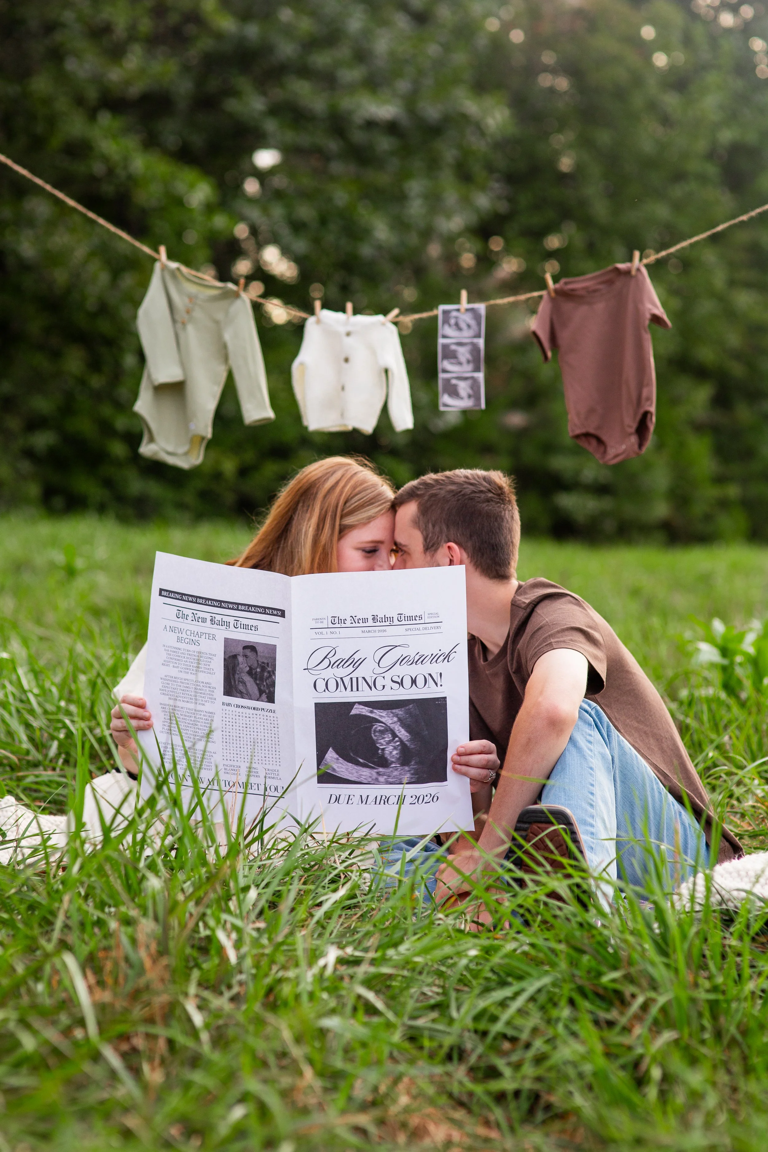 A couple sitting on grass, holding a newspaper with ultrasound images and an announcement about a baby born in March 2026. There are baby clothes hanging on a clothesline above them, and trees in the background.