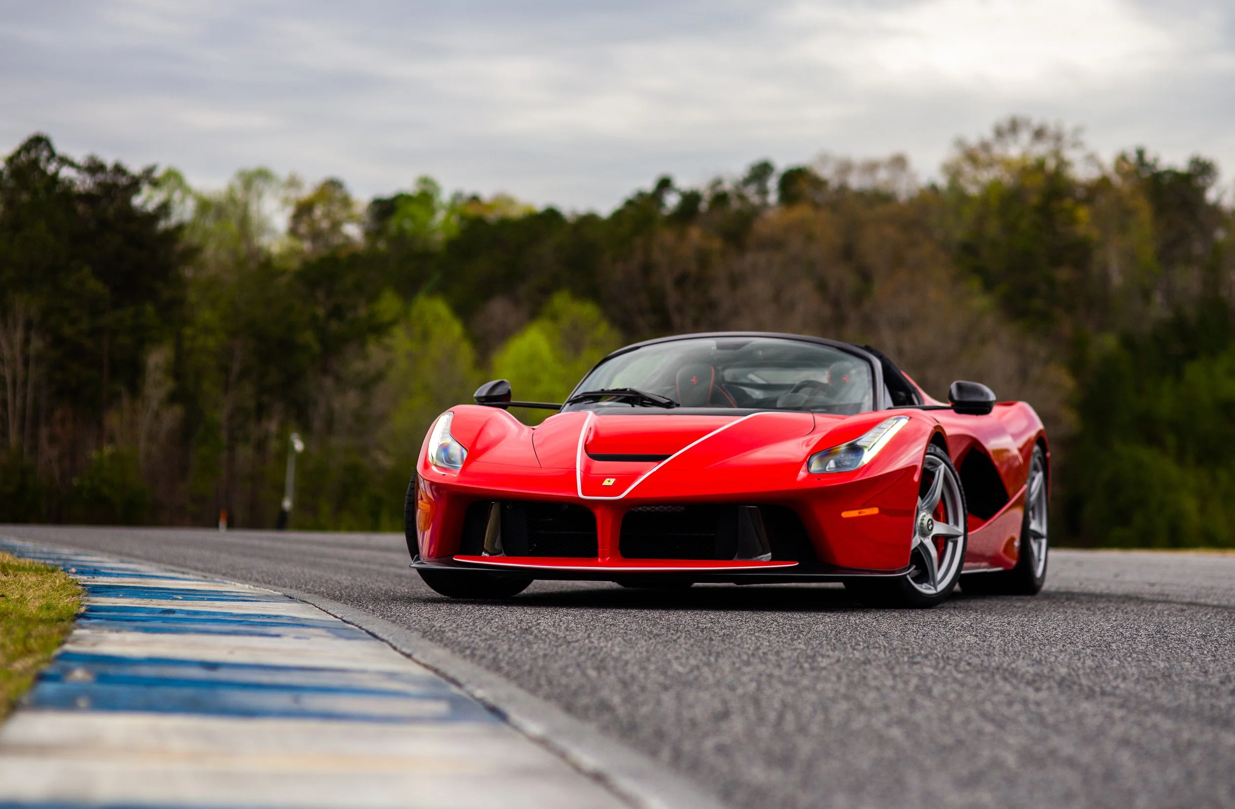 Red sports car on a race track with trees and cloudy sky in the background.
