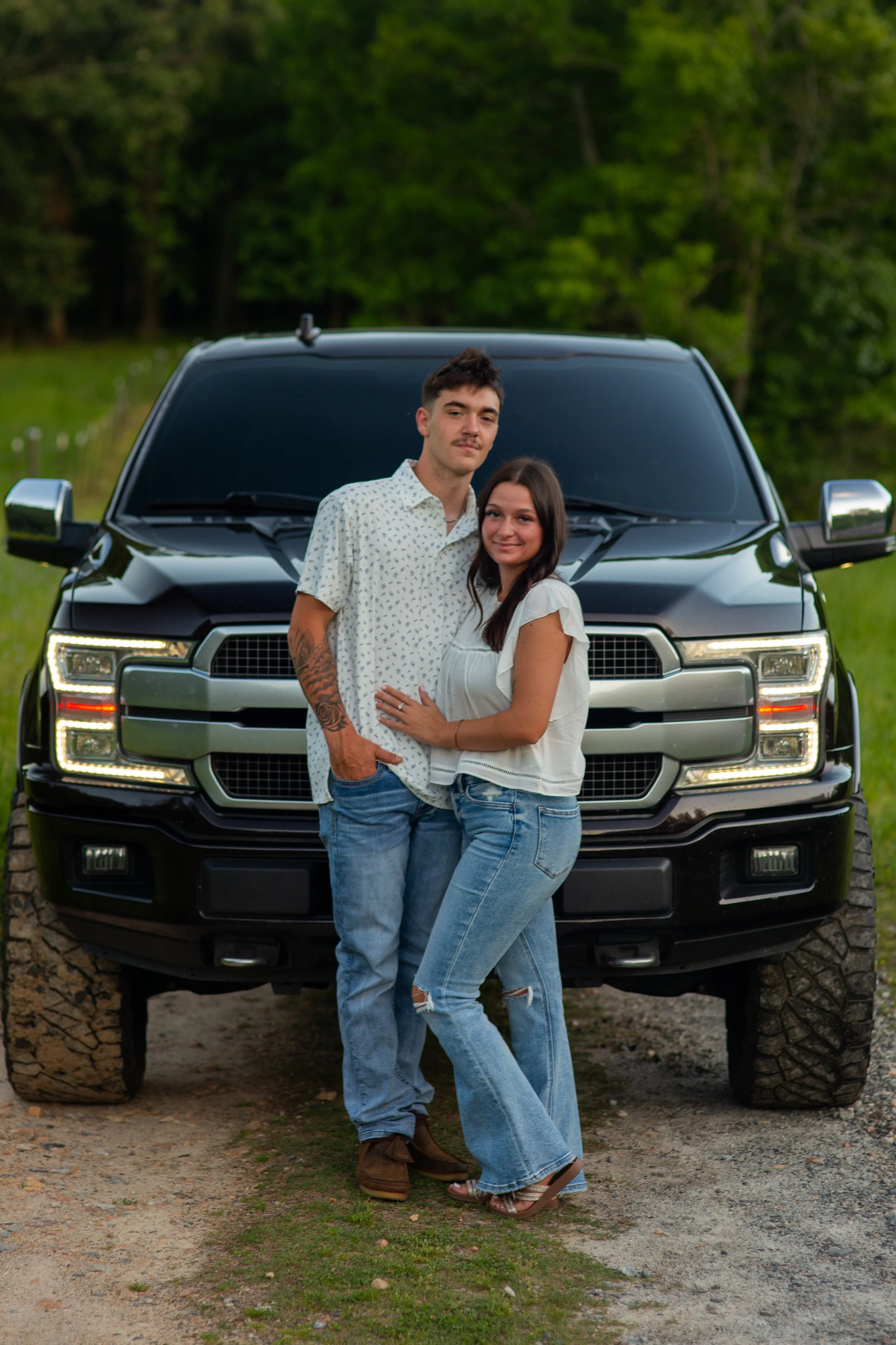 Young couple standing closely in front of a large black SUV on a dirt and grass trail in a wooded area. The man has short dark hair, a light mustache, and is wearing a white shirt with small patterns, ripped jeans, and brown shoes. The woman has long