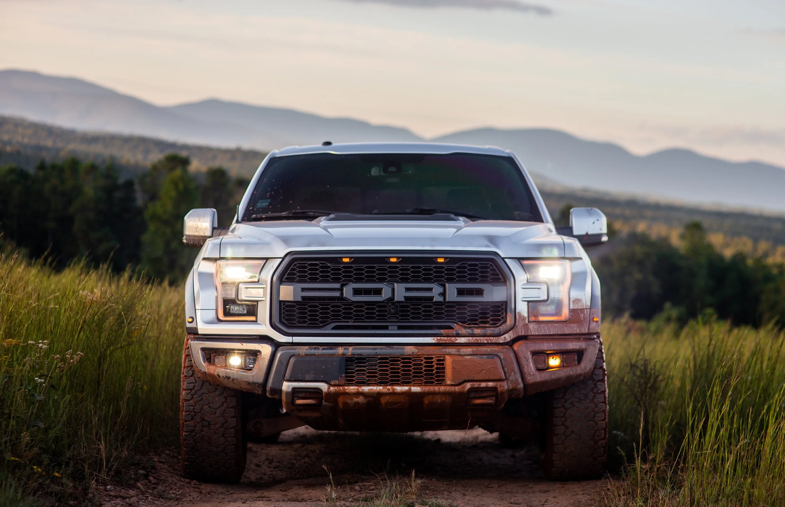 Front view of a silver Ford truck with mud-covered tires parked on dirt in a grassy field with mountains in the background during sunset.