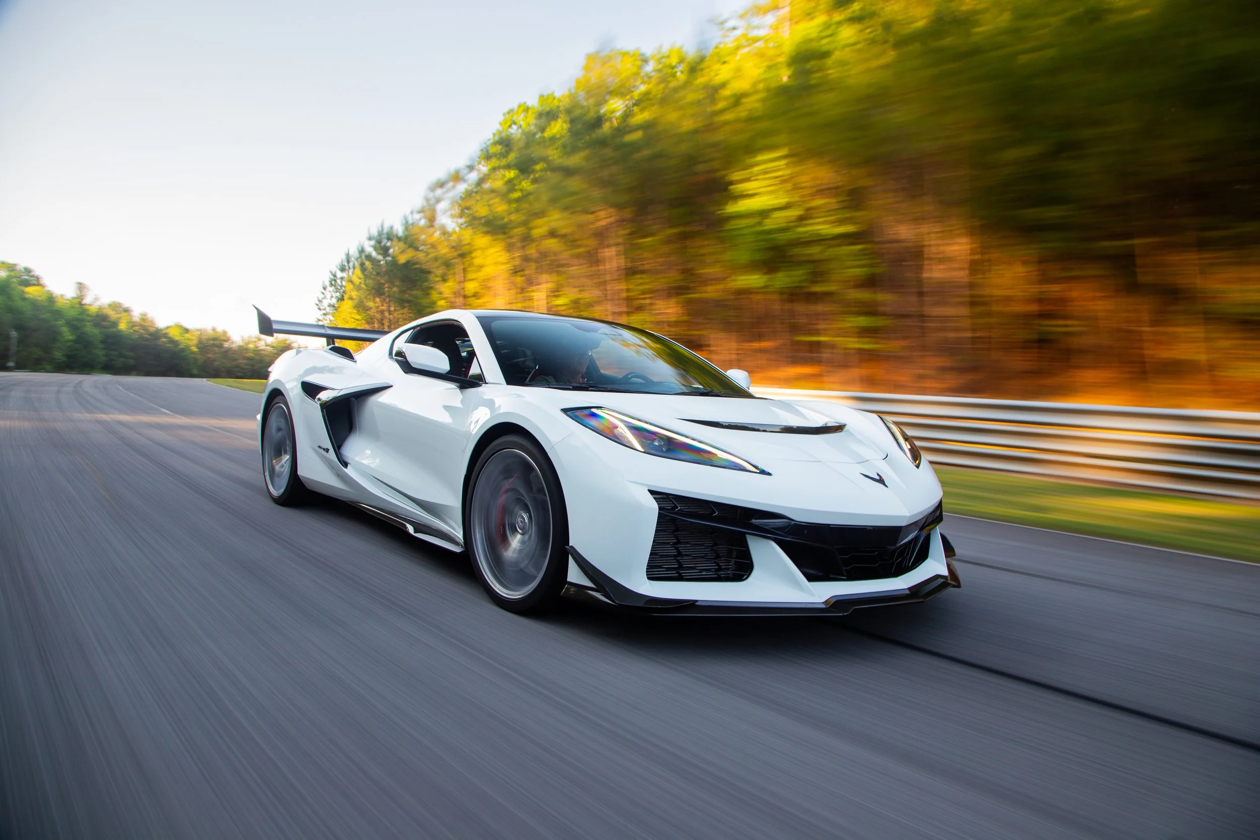 A white sports car, likely a Lamborghini, is speeding along a curvy road surrounded by trees with autumn foliage. The car has a sleek, aerodynamic design with a large rear wing.