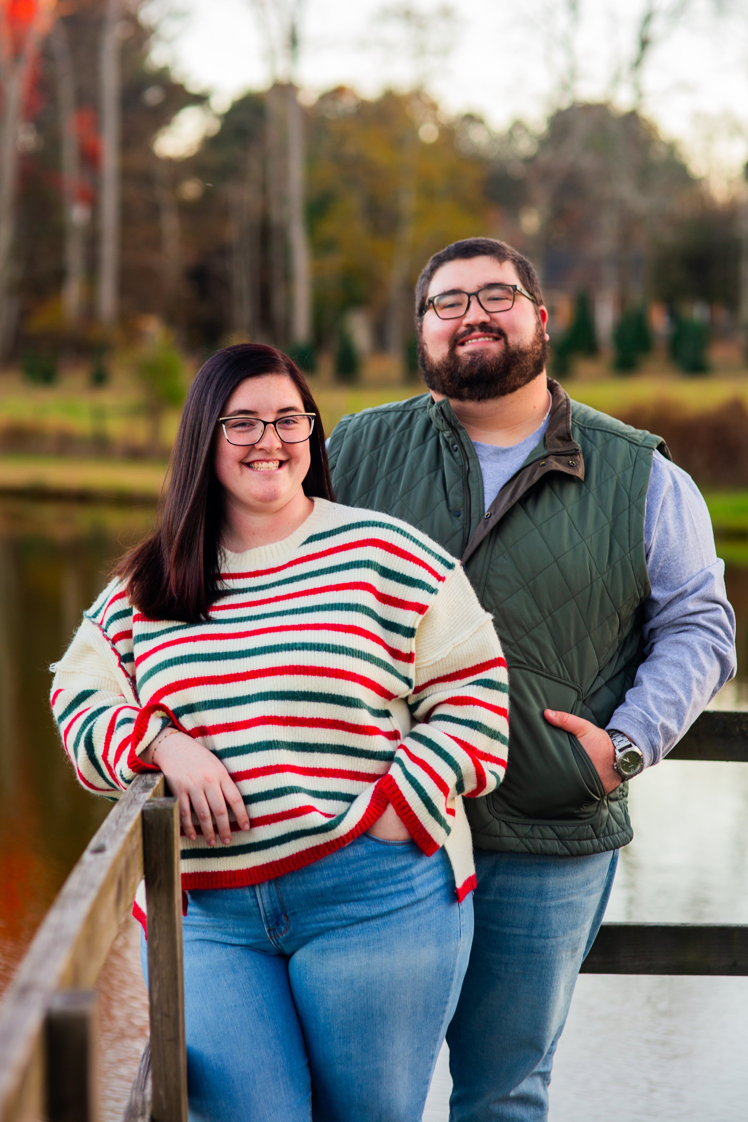 A smiling woman with long dark hair and glasses standing next to a smiling man with glasses and a beard, on a wooden bridge by a body of water with trees in the background.