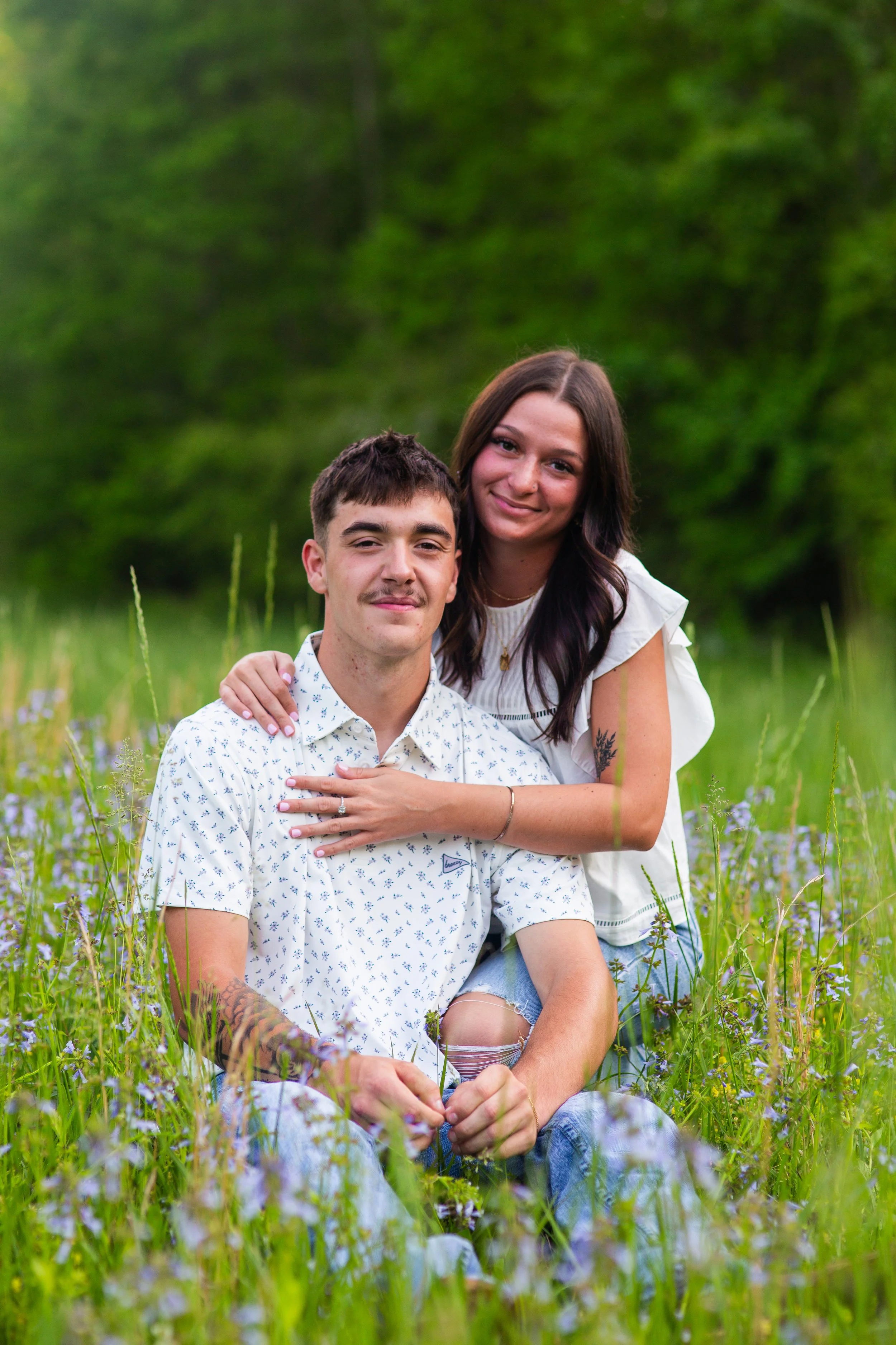 A young man and woman sitting and standing in a grassy field with purple flowers, smiling at the camera with lush green trees in the background.