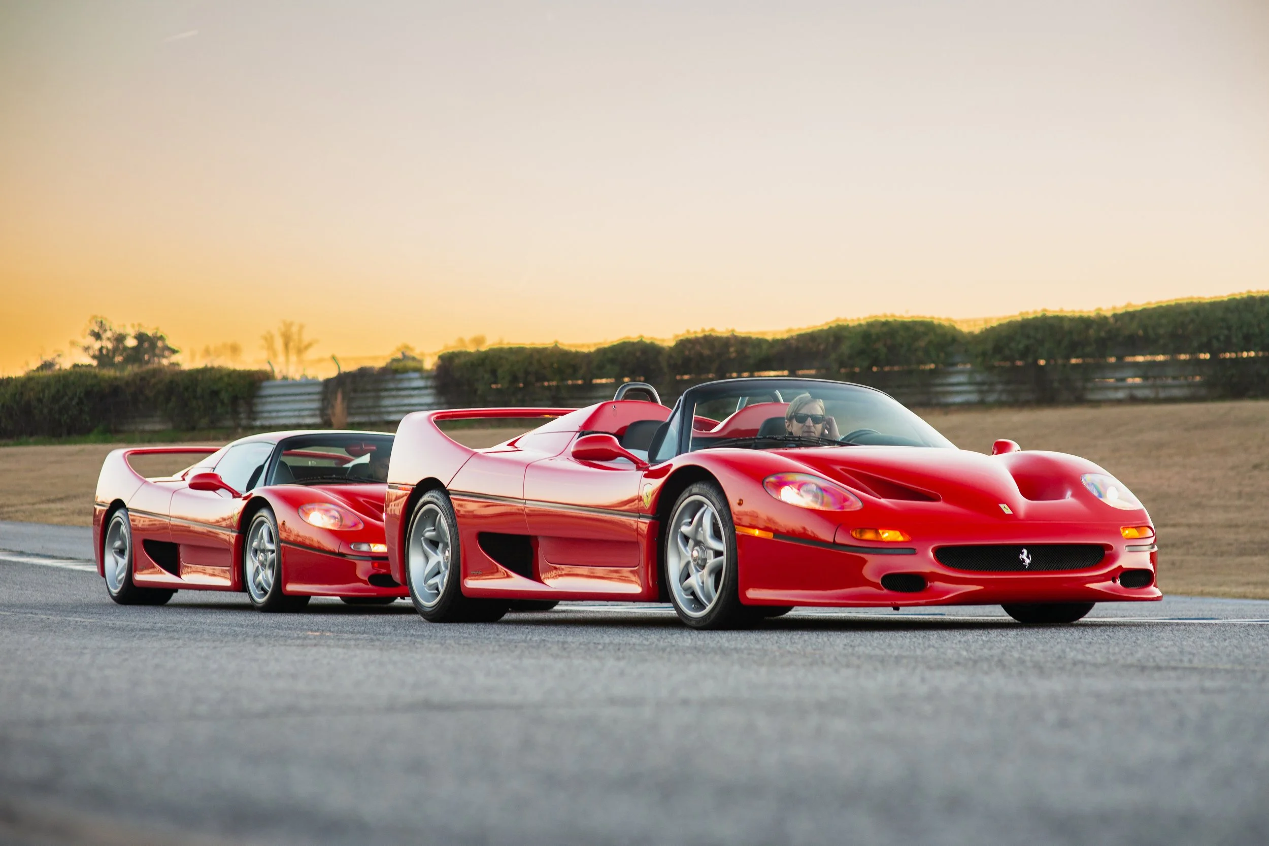Two red sports cars, a Ferrari and a Porsche, are driving side by side on a race track during sunset, with a man wearing sunglasses in the Ferrari.
