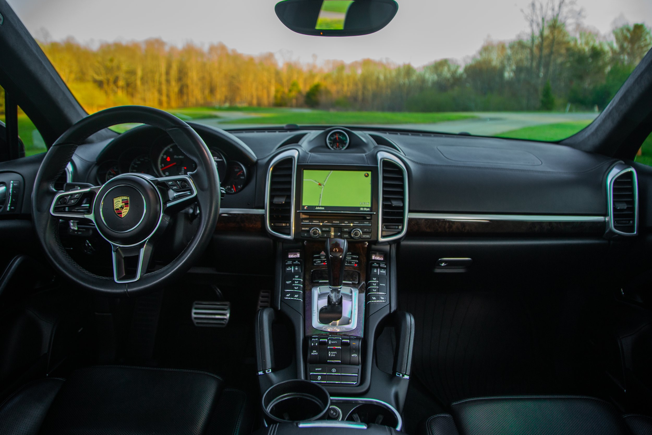 Interior view of a luxury car dashboard with a steering wheel featuring Porsche emblem, digital navigation screen, and gear shift, with a scenic countryside visible through the windshield.