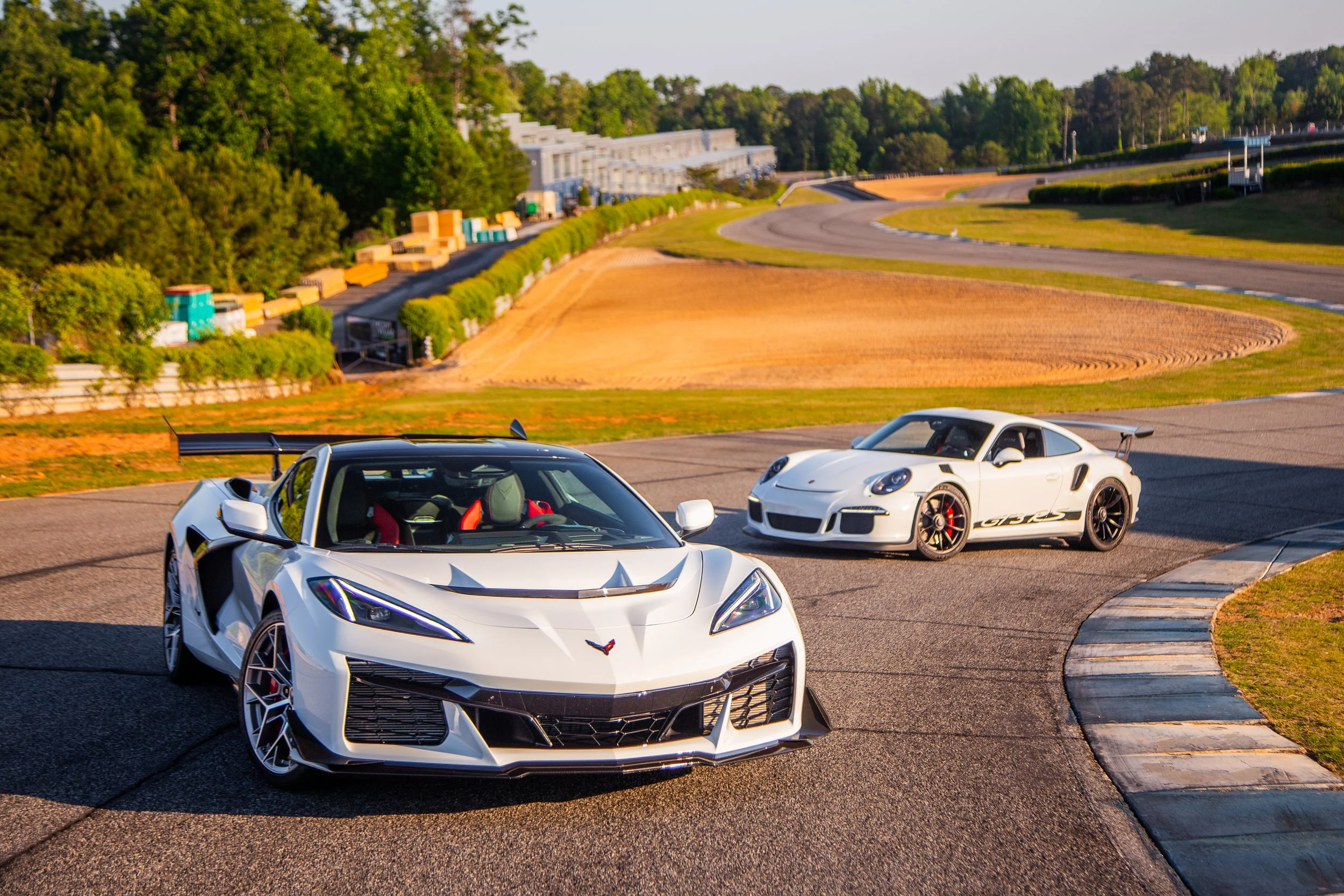 Two white sports cars, a Chevrolet Corvette and a Porsche 911 GT3 RS, are on a race track surrounded by green trees and hills. The cars are positioned next to each other on a curve of the race track.