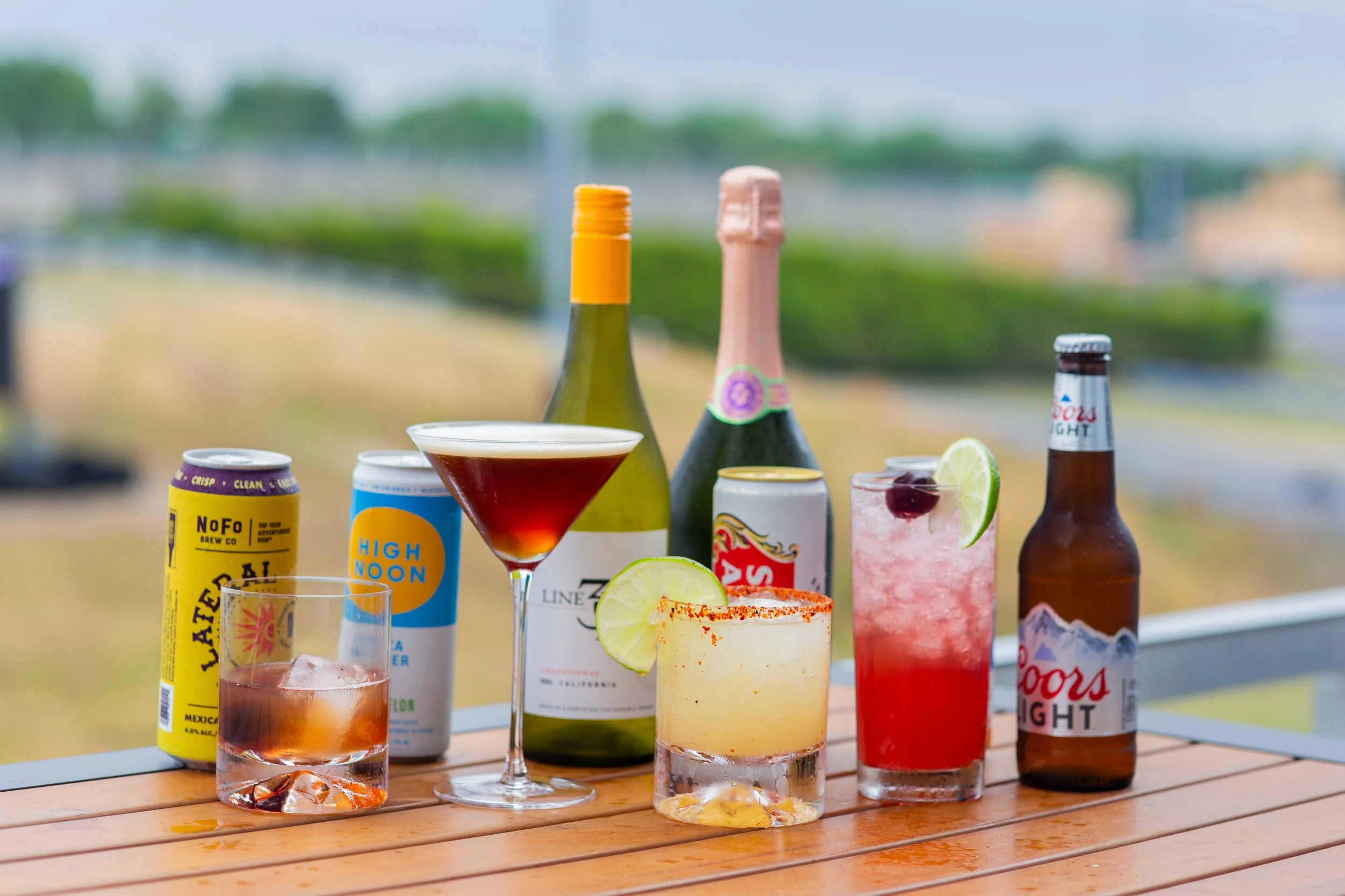 Assorted drinks and bottles on a wooden table, with a blurred outdoor background of trees and sky.