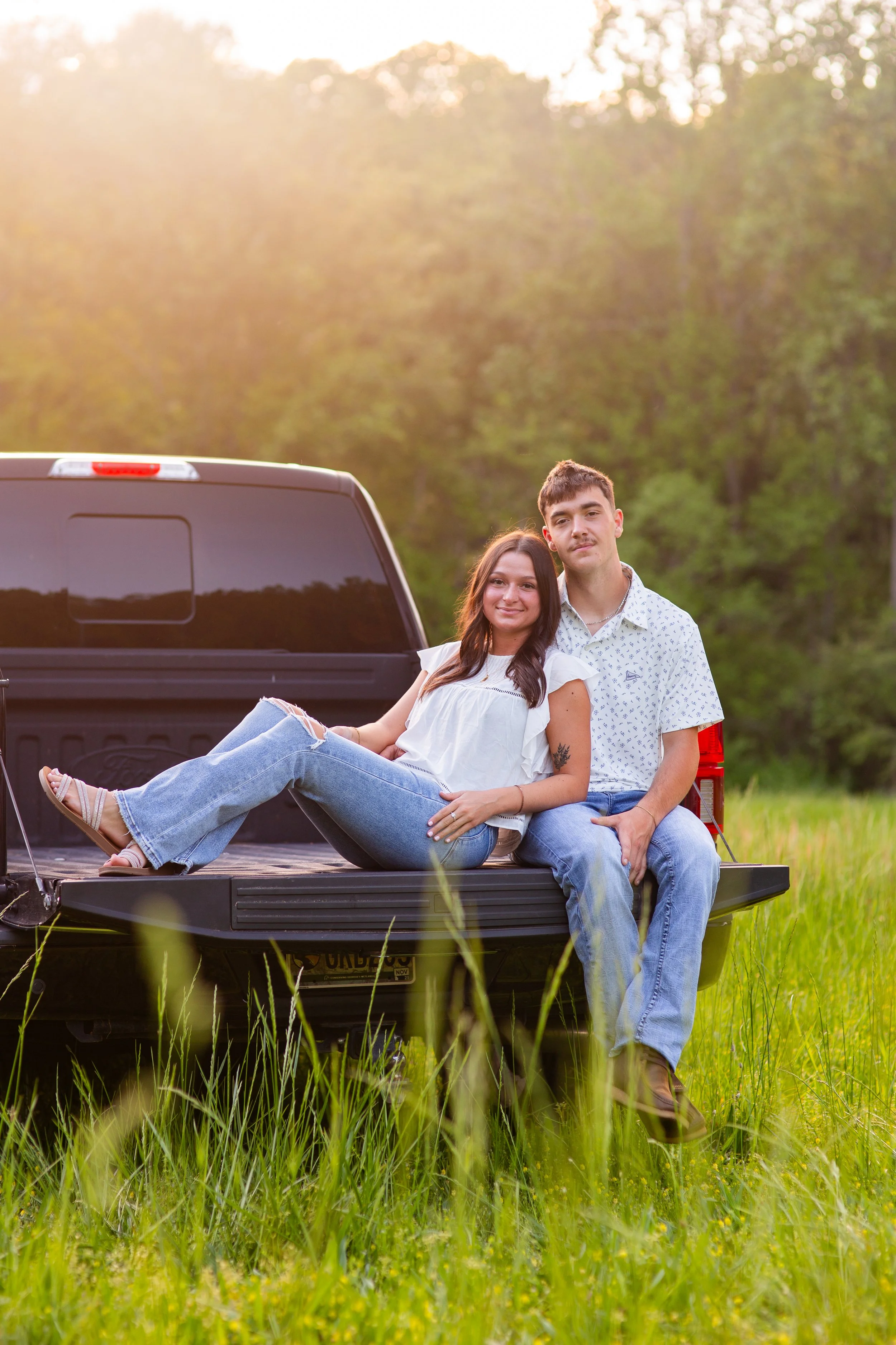 A young couple sitting on the tailgate of a black pickup truck in a green field during sunset, with trees in the background.