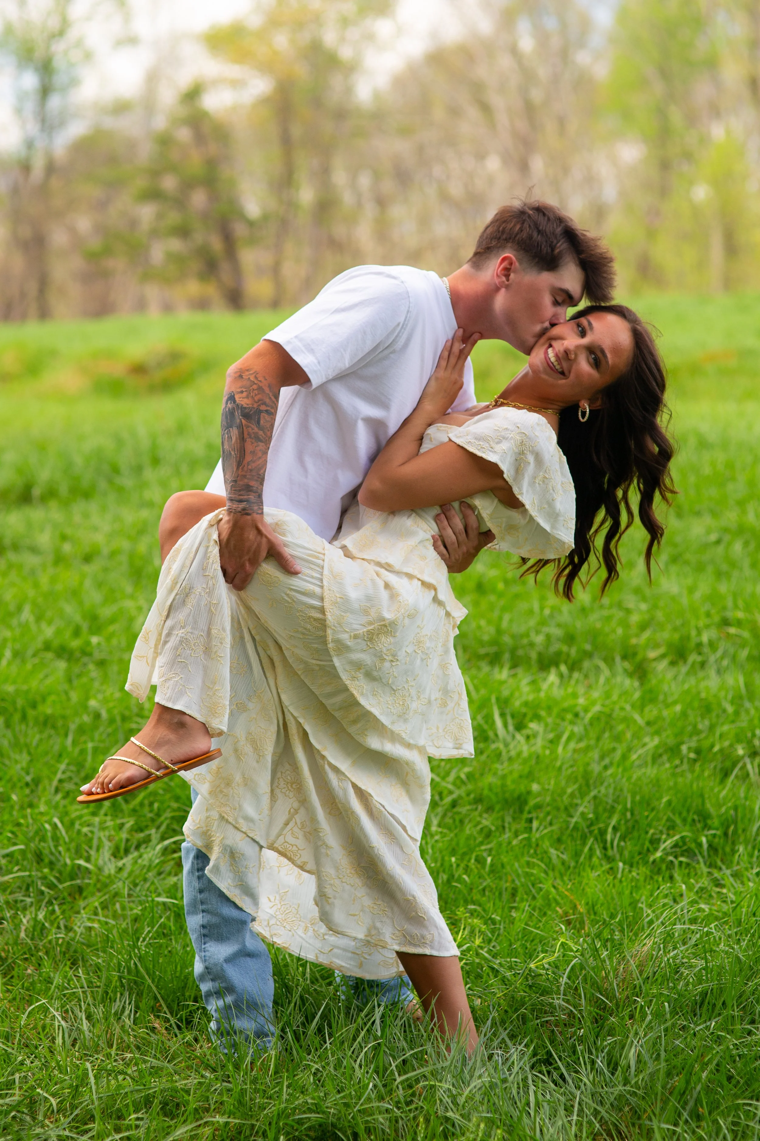 A young man in a white T-shirt is holding a smiling young woman in a yellow dress as she leans back in a grassy field. The man is kissing the woman's cheek. The background features a blurred natural setting with trees and grass.
