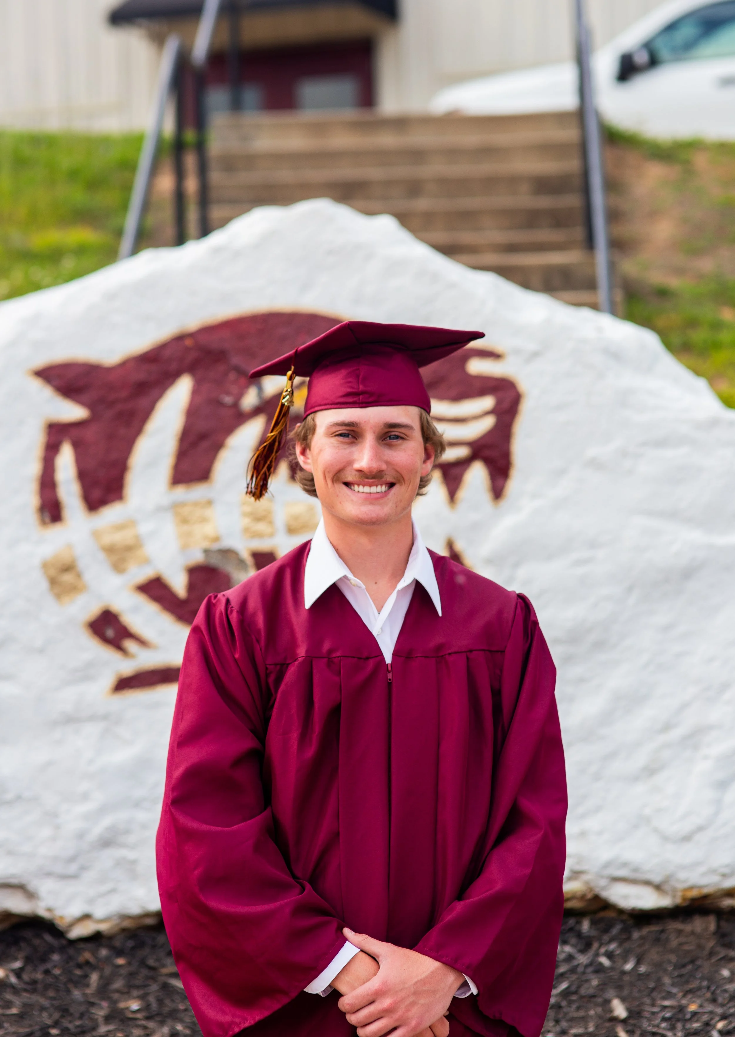 A young man in a maroon graduation gown and cap stands outdoors in front of a rock with a maroon dinosaur logo painted on it, smiling. Behind him are stairs, a building, a parked white car, and green foliage.