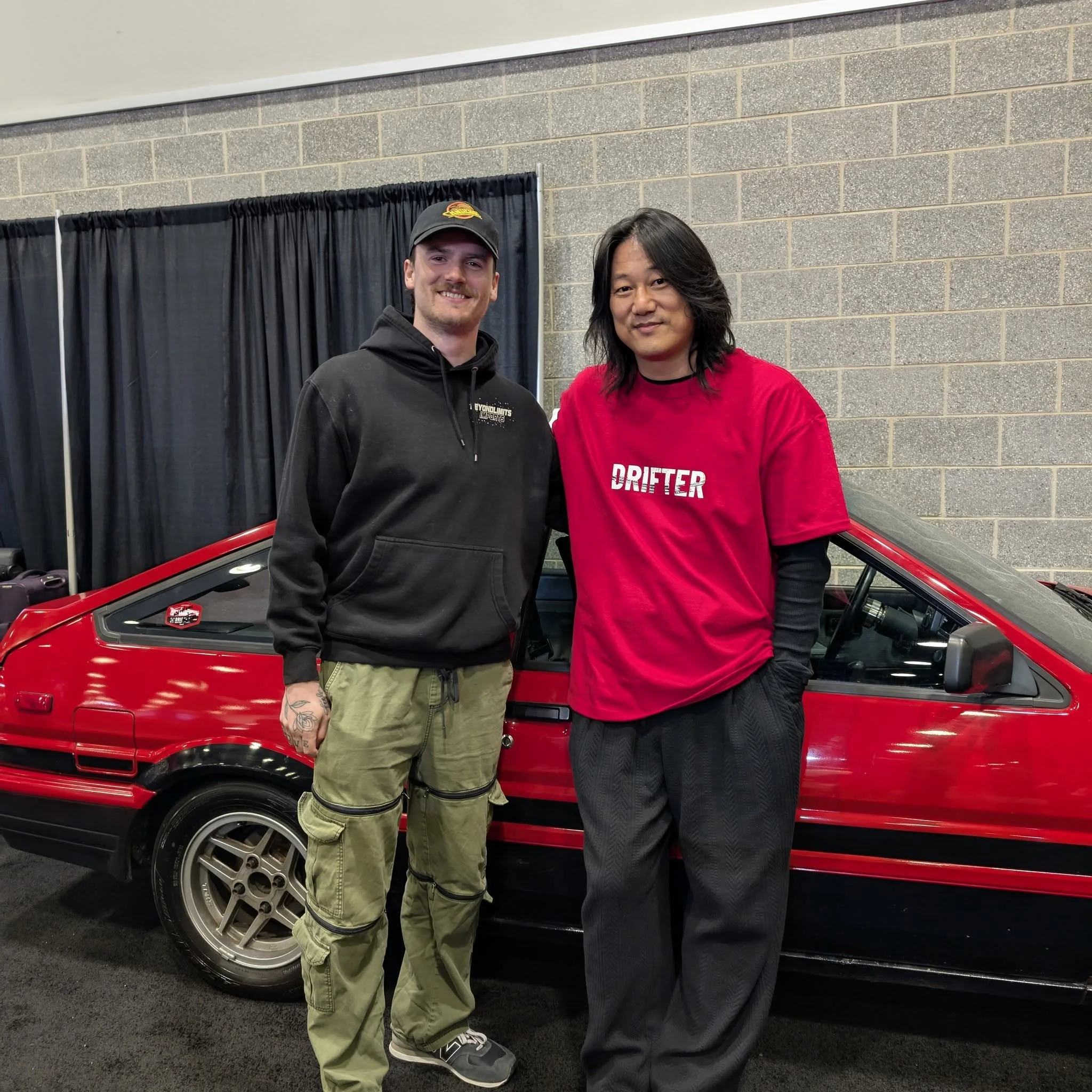 Two men standing next to a red sports car indoors against a gray brick wall. One man is wearing a black hoodie and khaki pants, and the other is in a red shirt and black pants. They are smiling and posing for the photo.