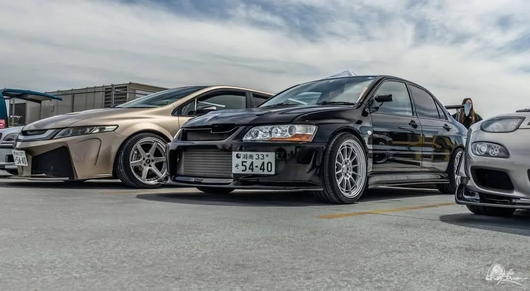 A black modified sports sedan parked between a beige sedan and a silver sports car in an outdoor parking lot.