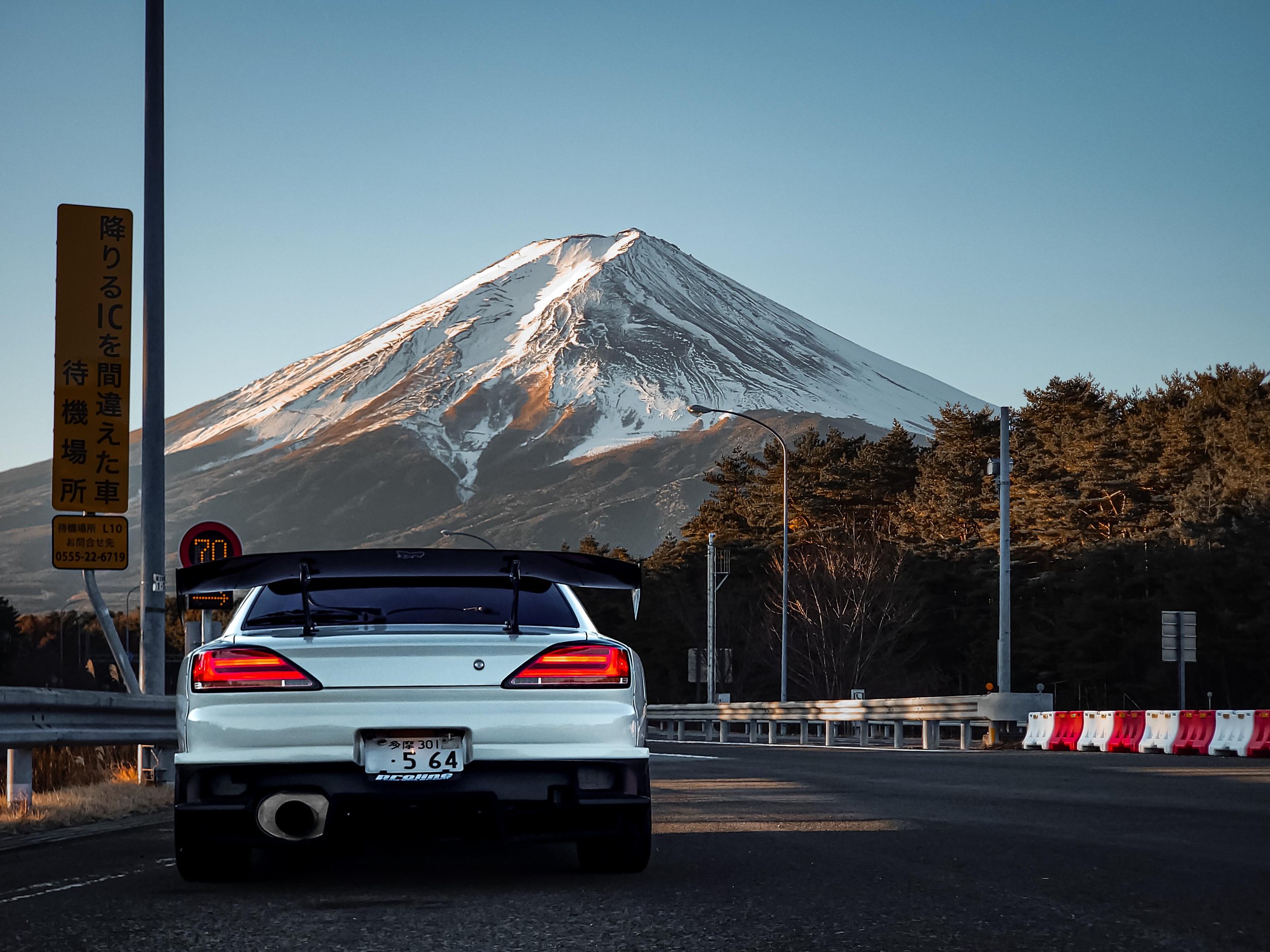 Rear of a white car stopped at a toll booth with mountains, including Mount Fuji snow-capped peak, in the background.