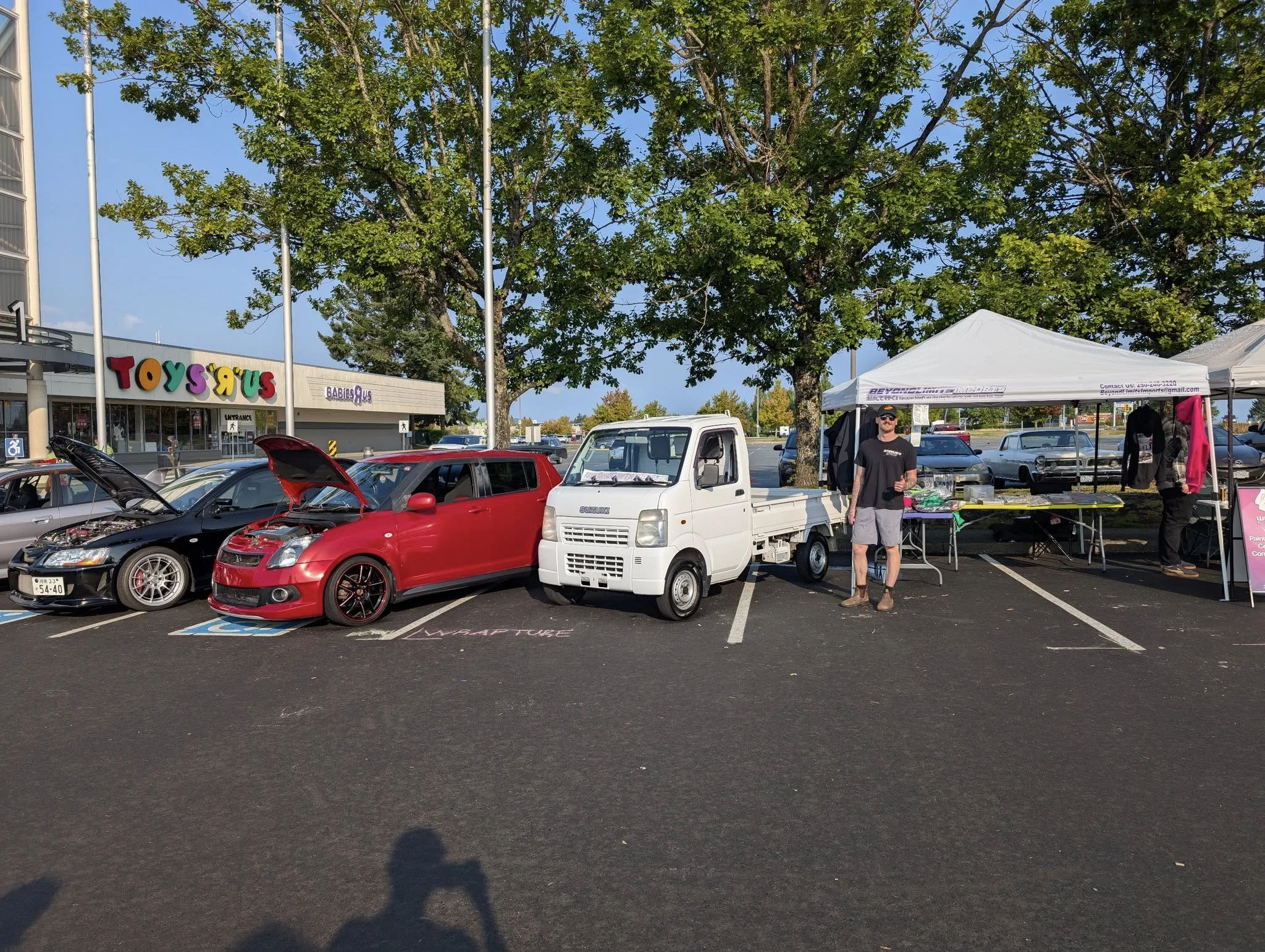 Parking lot with cars, including a red sports car and a white mini truck, near a Toys R Us store. A man stands under a white tent with a table and hanging clothes, and there are large trees and a clear blue sky.