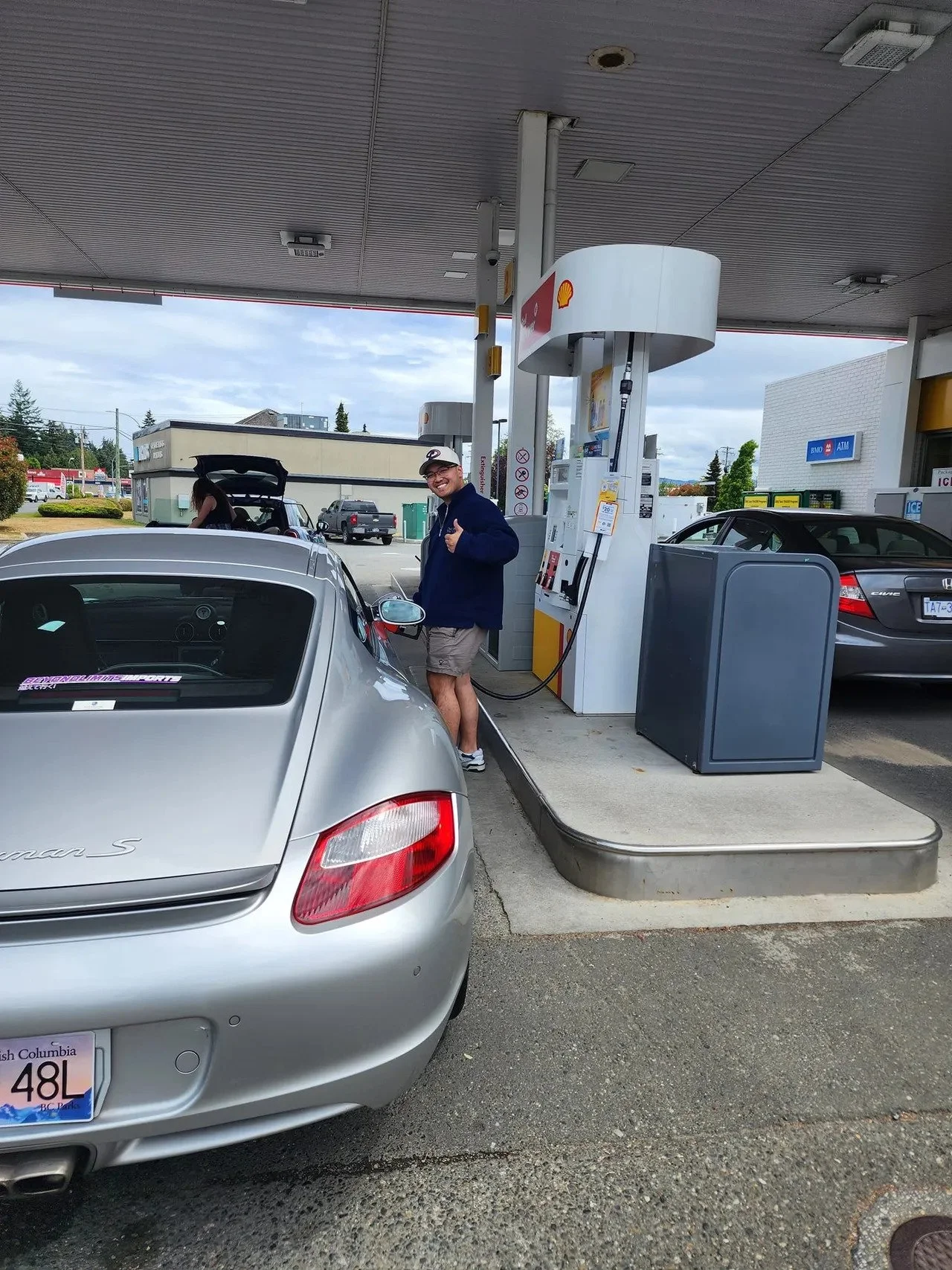 A man standing next to a silver Porsche Cayman S at a Shell gas station, giving a thumbs up and smiling, with the gas pump nearby.