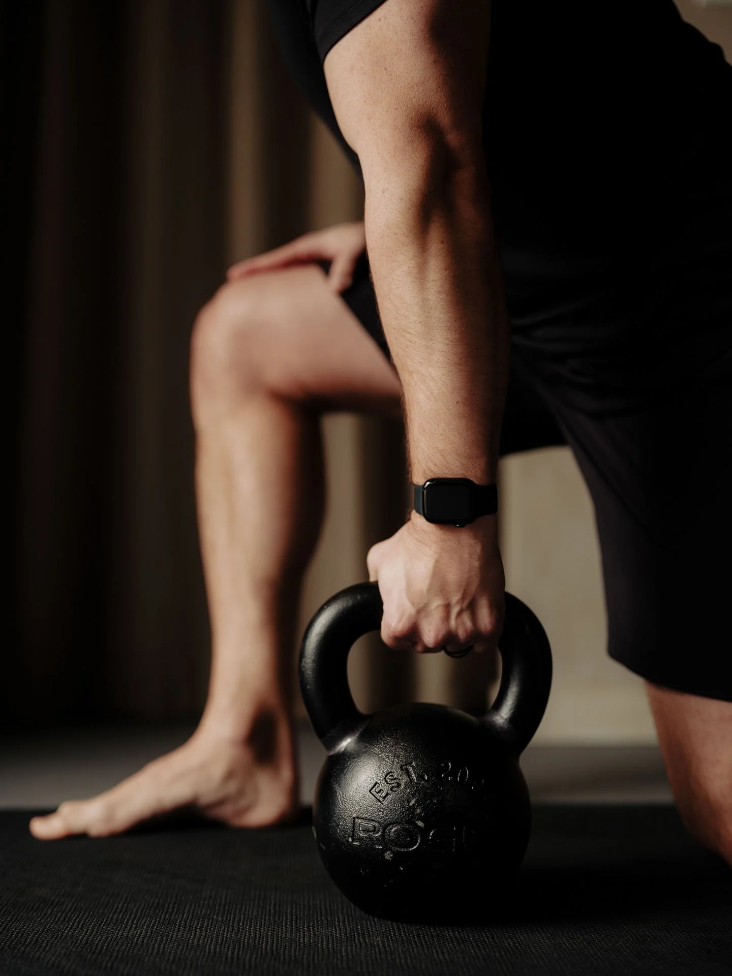 Close-up of a person holding a kettlebell while performing a workout, wearing a smartwatch, with their other hand and foot visible on a black workout mat.