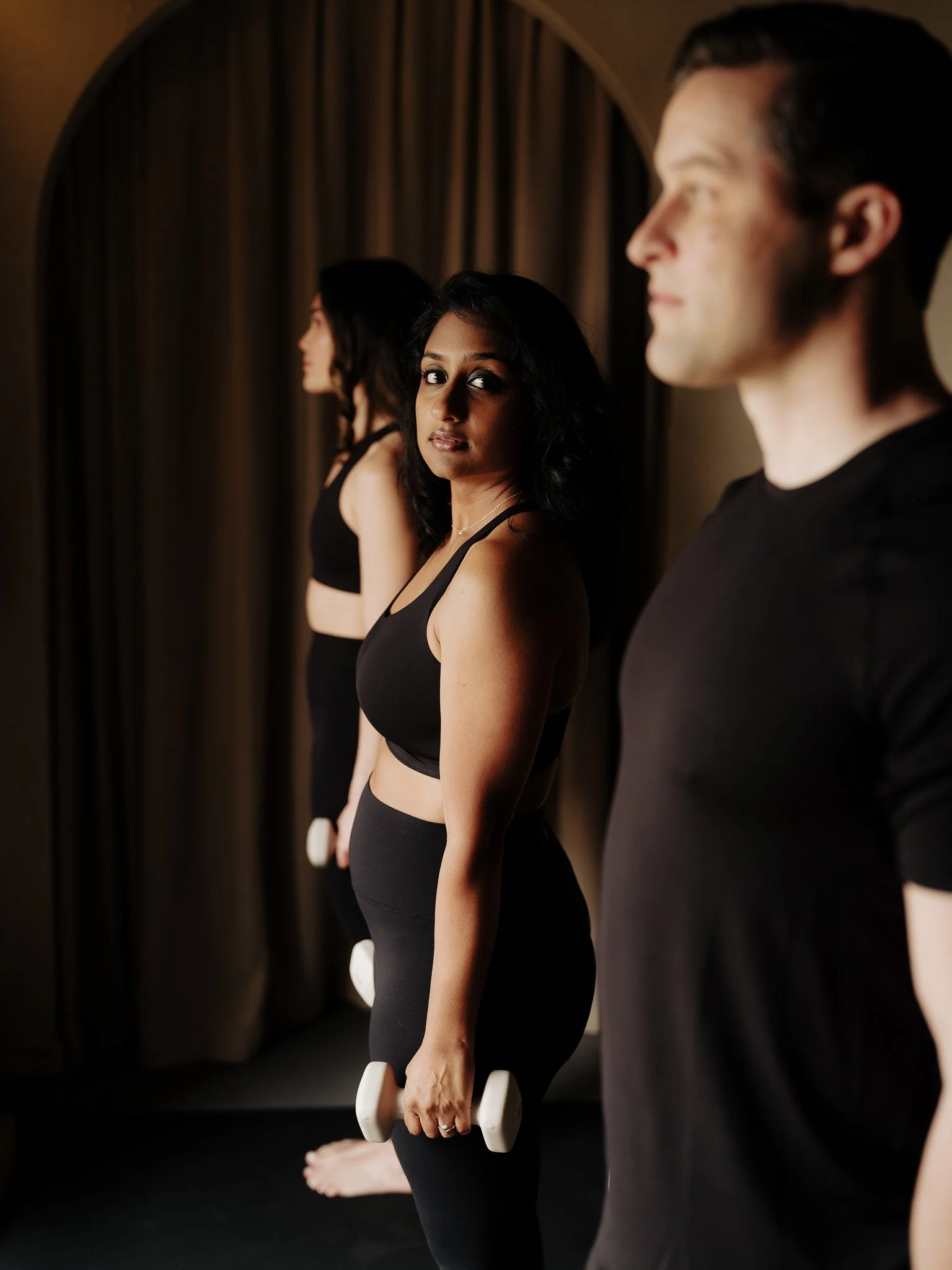Three women practicing yoga, standing on a mat, holding white weights with a focused expression, facing a mirror with brown curtains in the background.