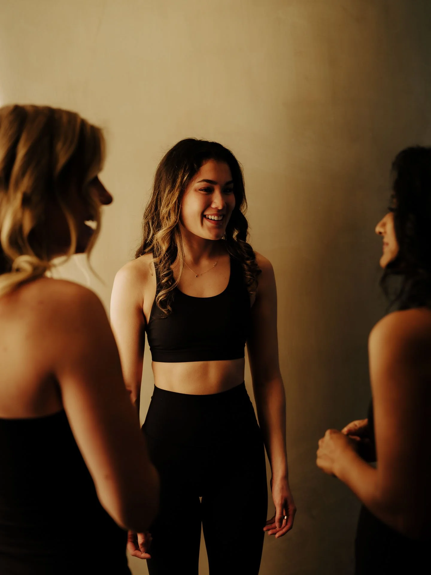 Three young women wearing black workout clothes, smiling and chatting in a dimly lit room.