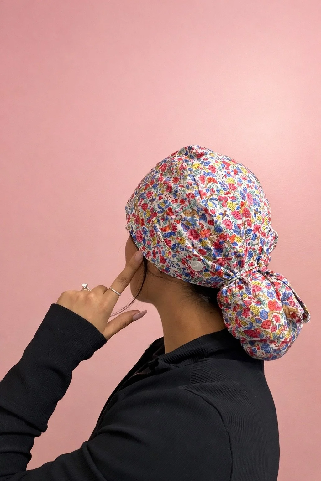 A woman wearing a colorful floral scrub cap, black top, pointing at her ear, against a pink background.