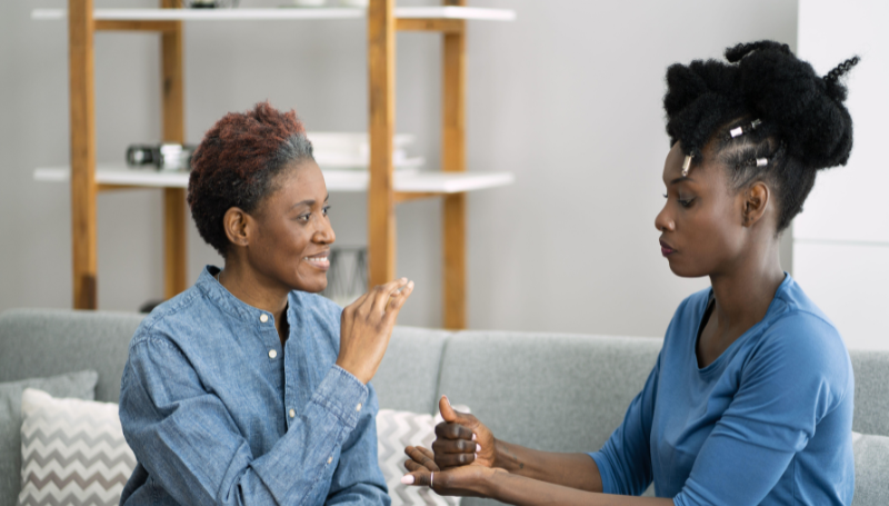 A woman with short curly hair smiling and signing with another woman with long braided hair sitting on a comfy couch.
