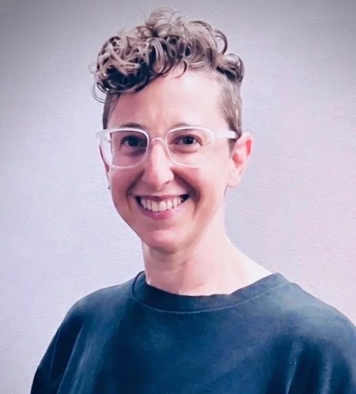Portrait of Lori Keslowitz, Founder, smiling with short, curly hair, wearing clear glasses and a dark shirt, posing against a plain wall.